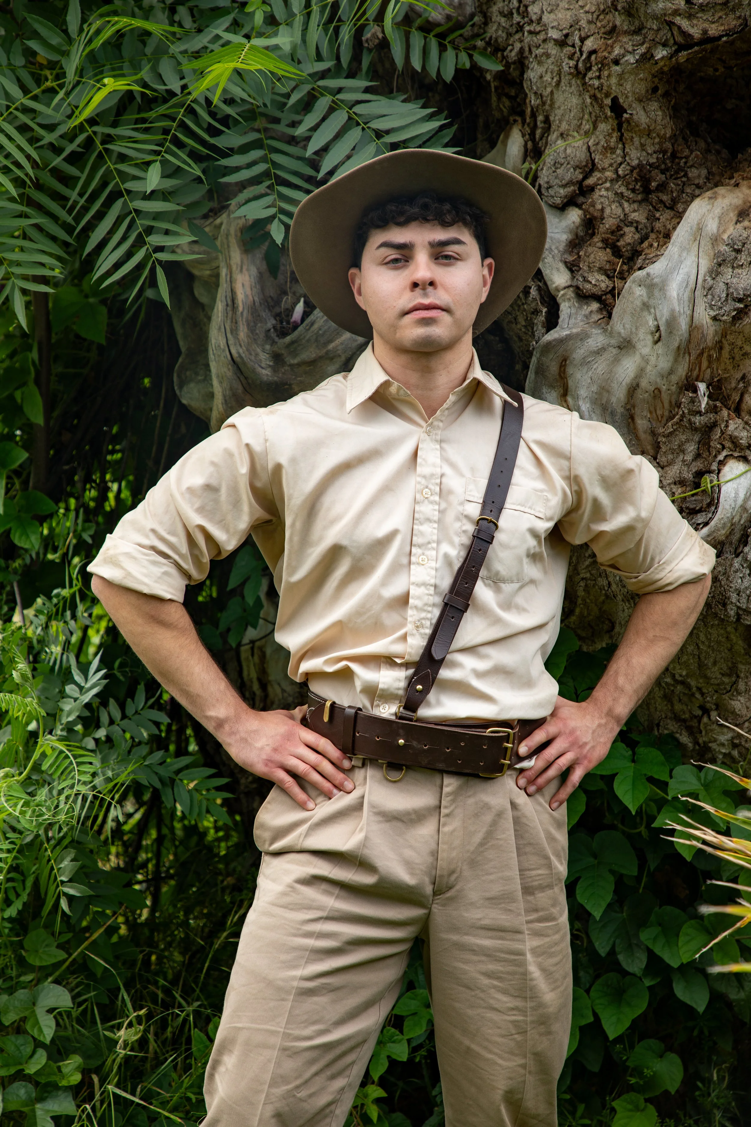Joey Chacho, dressed as a park ranger or explorer, wearing a beige shirt, beige pants, a wide-brimmed hat, and a leather belt, stands confidently with his hands on his hips in front of dense green foliage and a tree trunk.
