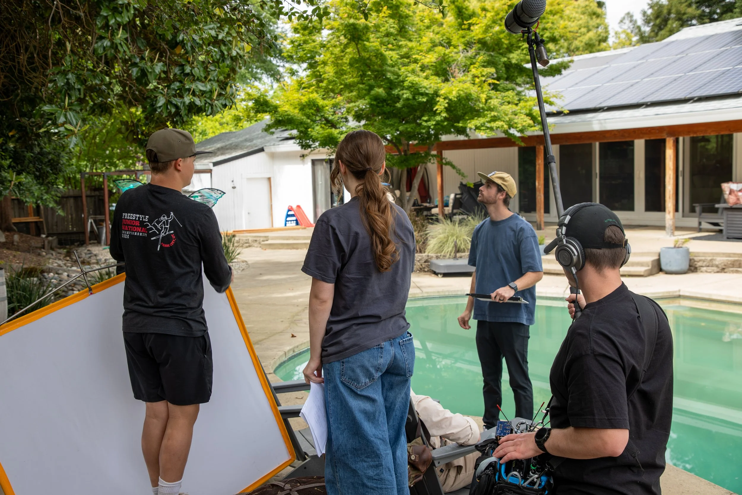 A film crew working outdoors near a swimming pool, with green trees in the background. The crew includes with butterfly wings on their back, another person holding a clipboard, and a person operating camera equipment, with a boom microphone overhead.