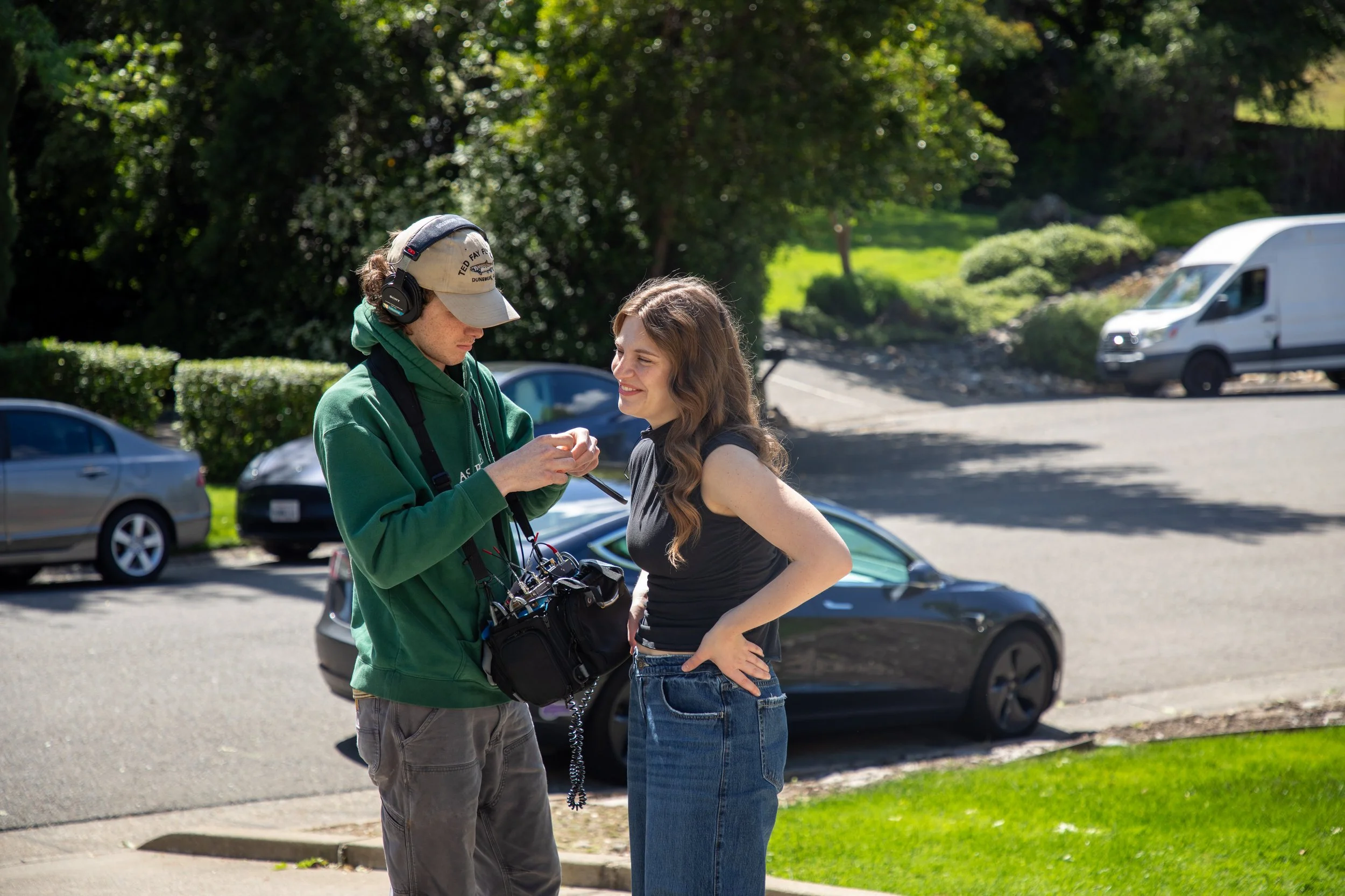 A member of the crew is putting a mic on an actor. It is sunny and lush outside.