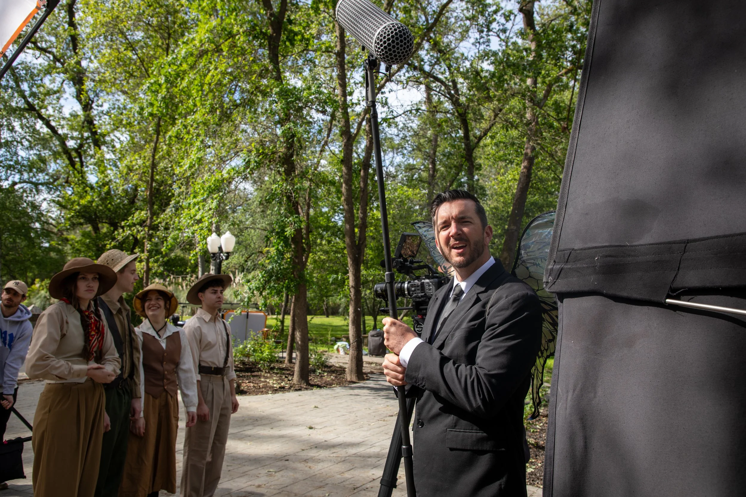 A man dressed in a black suit and tie is standing in front of a microphone set up outdoors. He is holding a piece of paper and appears to be speaking. Several young people dressed in tan shirts and hats are standing nearby. film equipmentalso visible