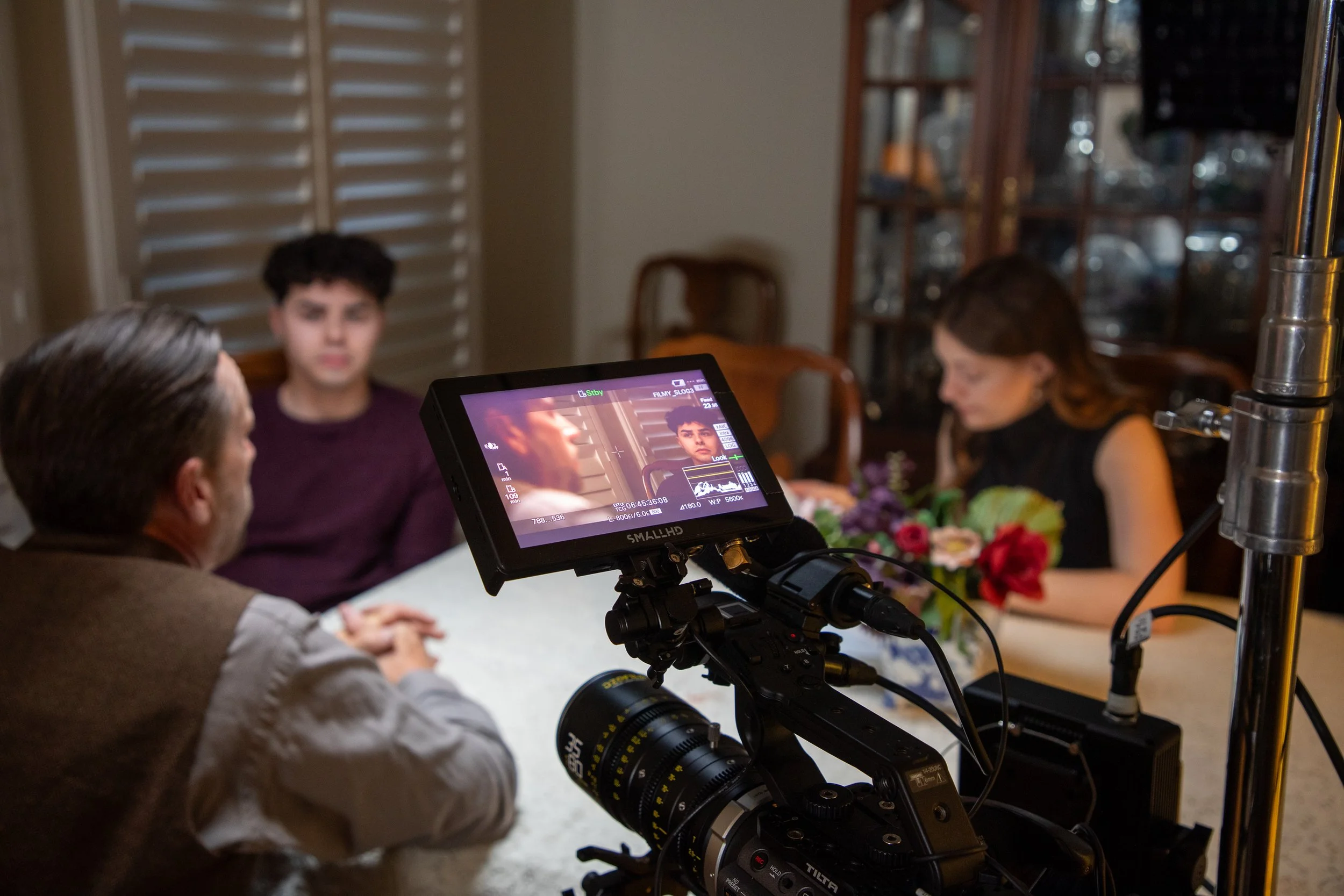 A film crew is shooting a scene indoors with three people visible: a woman, a young man with dark curly hair, and a man. The scene is being captured on a professional video monitor, and there is a bouquet of flowers on the table in front of them.