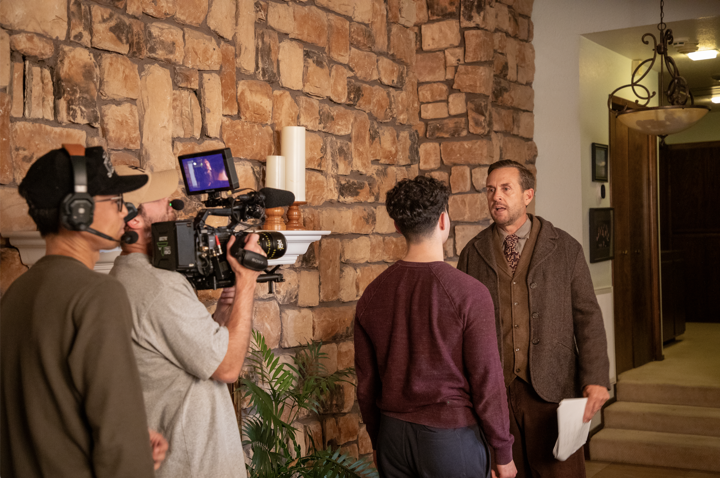 Filming crew recording a scene with two men in conversation near a stone fireplace, in a warm interior setting.