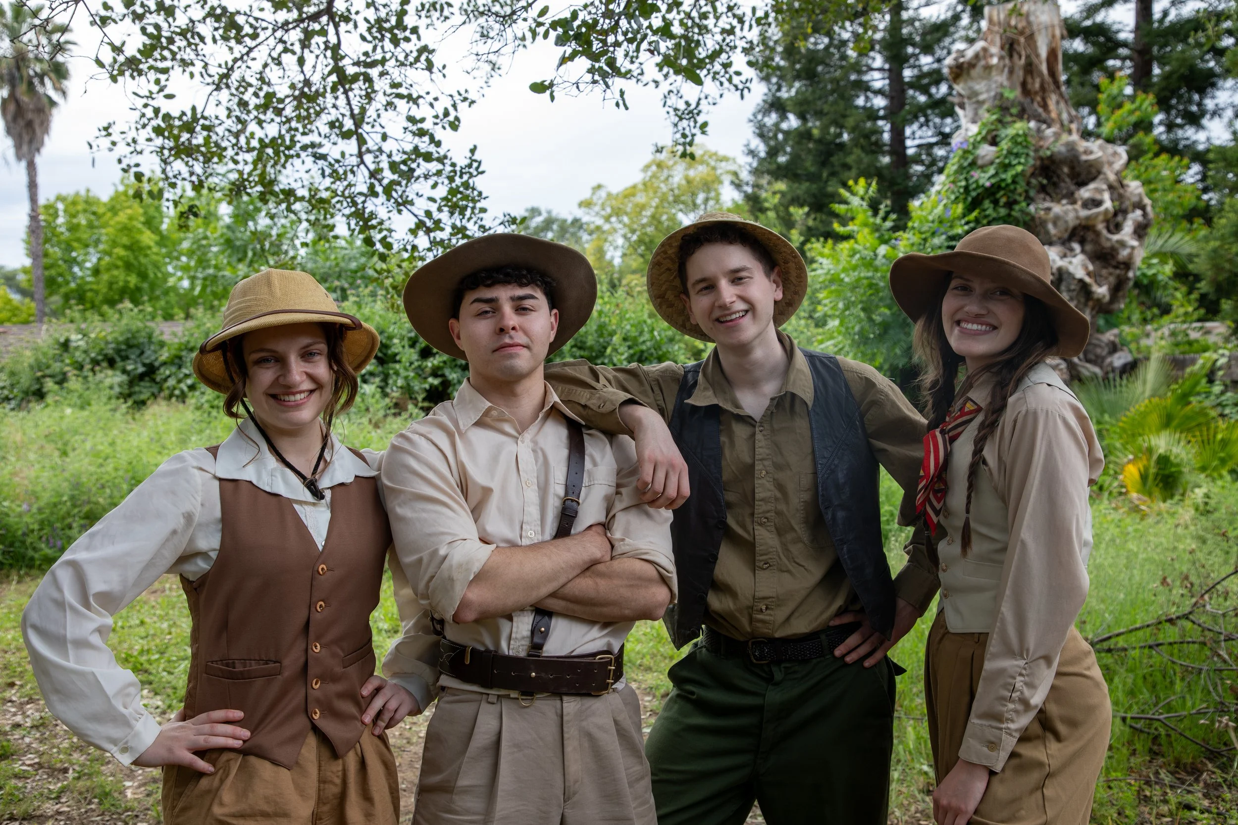 Four young adults dressed in safari or explorer outfits, smiling and standing outdoors in a lush, green, wooded area with trees and plants.