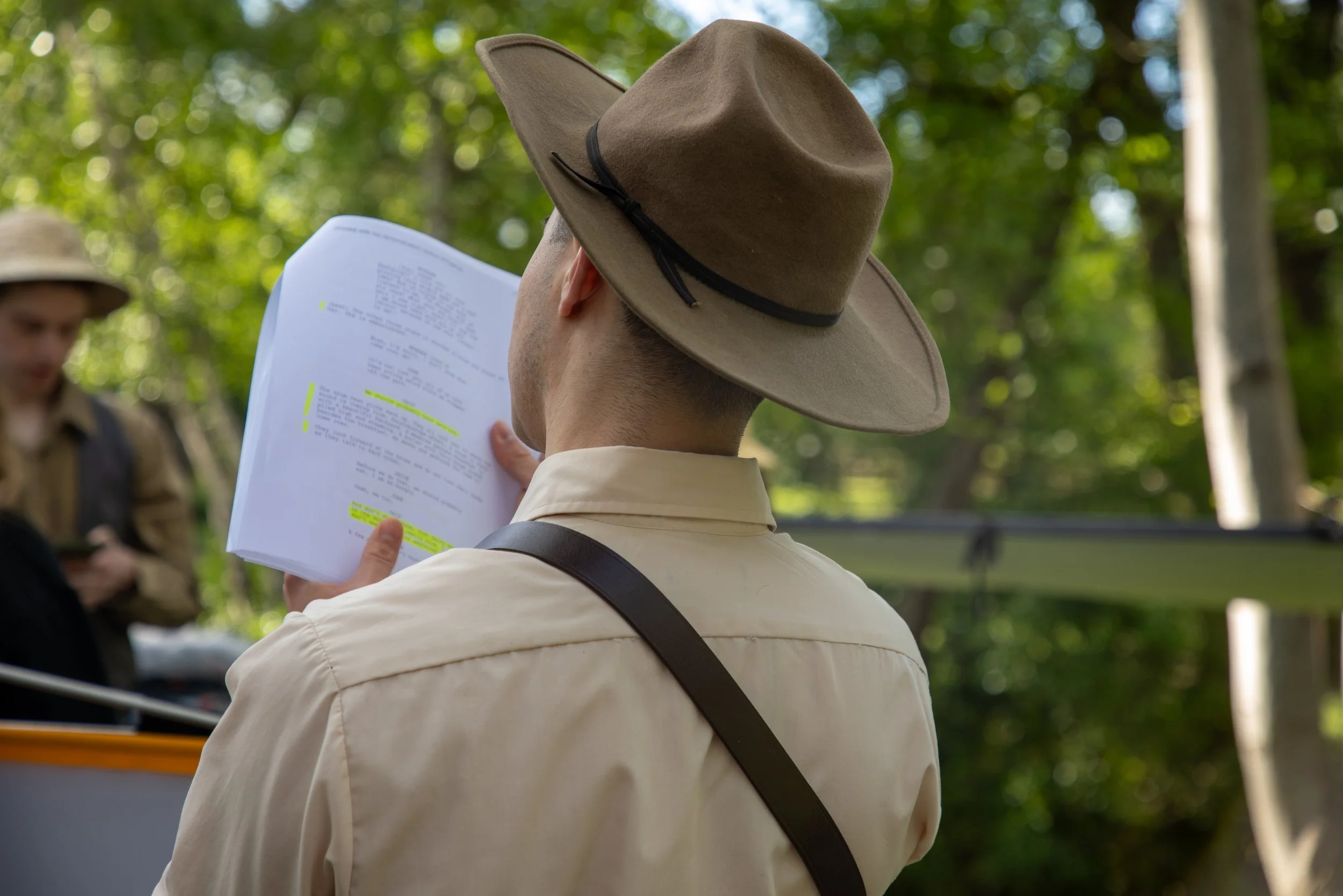 A person in a beige uniform and wide-brimmed beige hat is holding a script or screenplay with highlighted sections, standing outdoors among green trees. In the background, another person is partially visible, also dressed in a uniform and wide-brimme