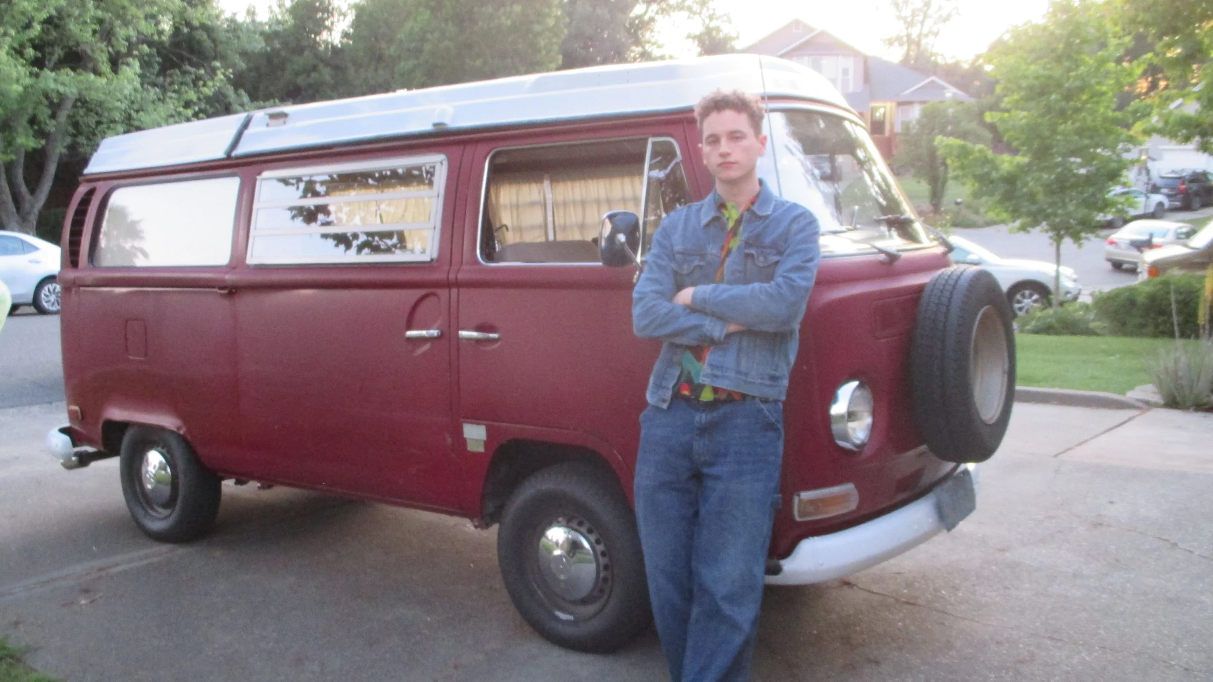 A young man with crossed arms standing in front of a vintage red camper van, with a background of trees and a residential neighborhood at sunset.