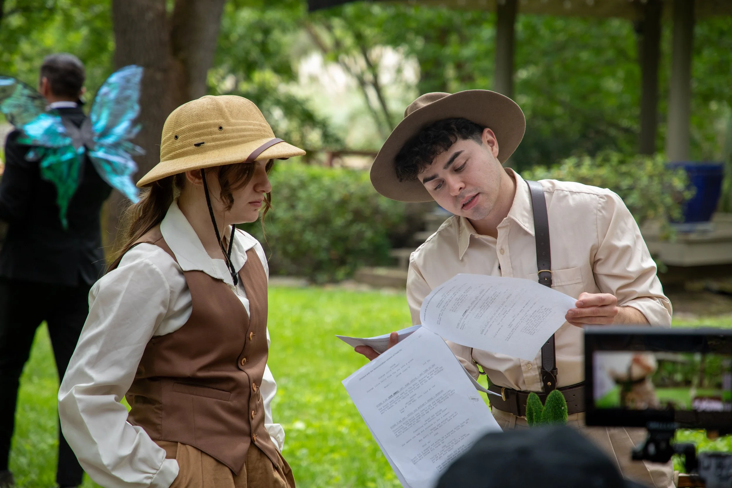 Two actors dressed in safari gear review scripts outdoors in a space with green trees and grass.