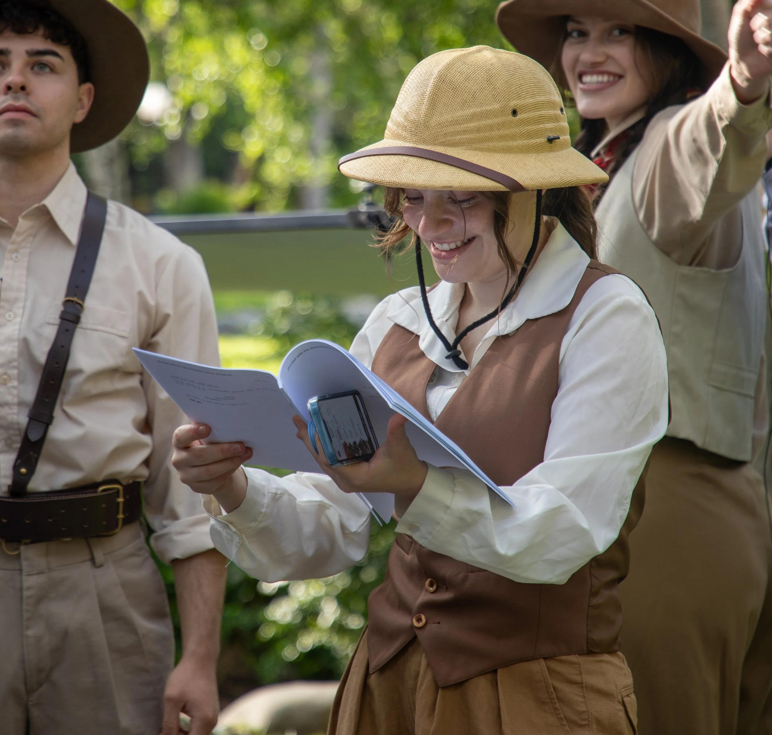 Three children dressed as explorers, wearing safari hats and uniforms, standing outdoors in a natural environment with trees in the background. One girl is smiling and holding a booklet.