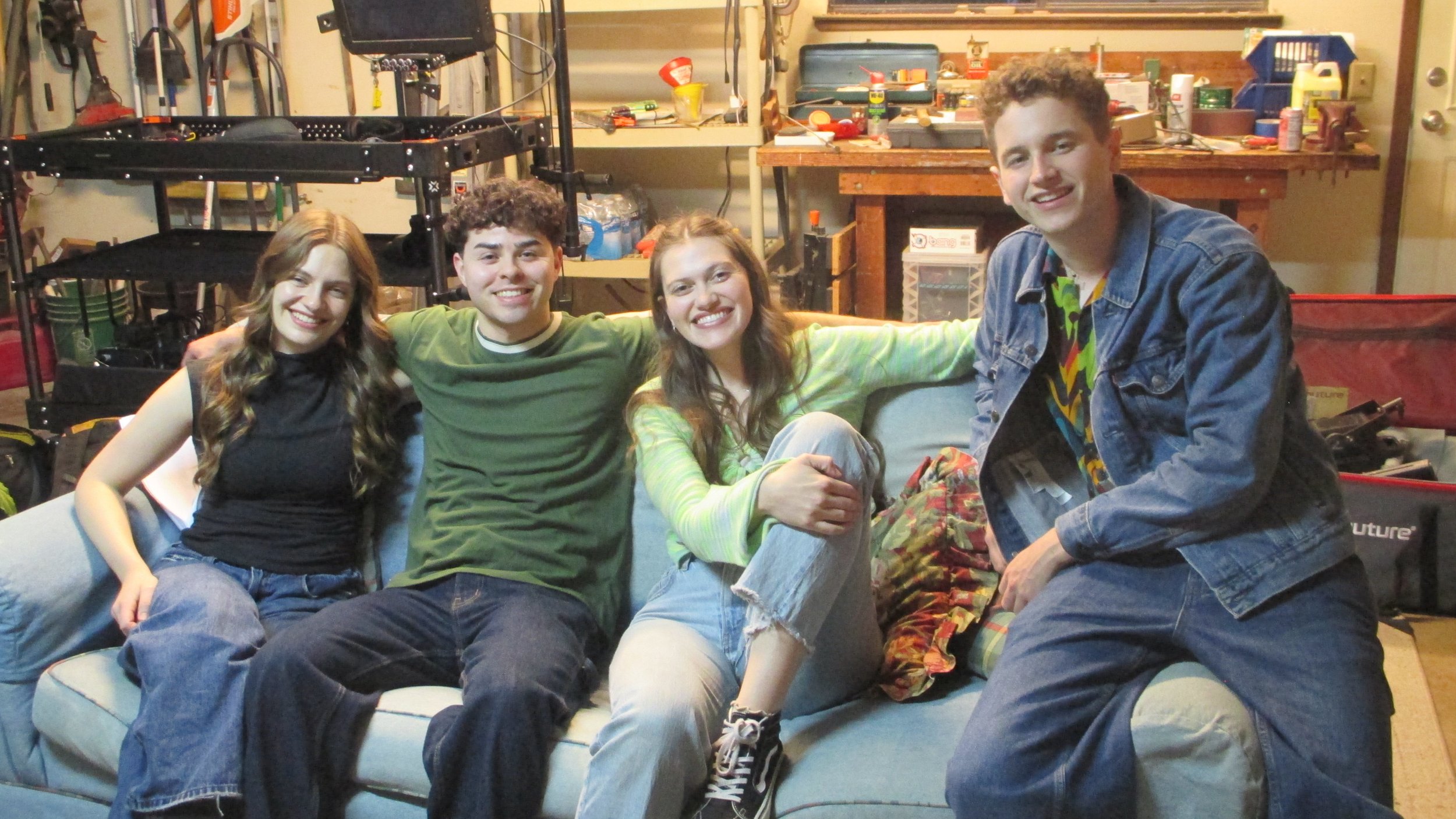 Four young people sitting together on a couch in a cluttered garage. They are smiling and appear to be friends.
