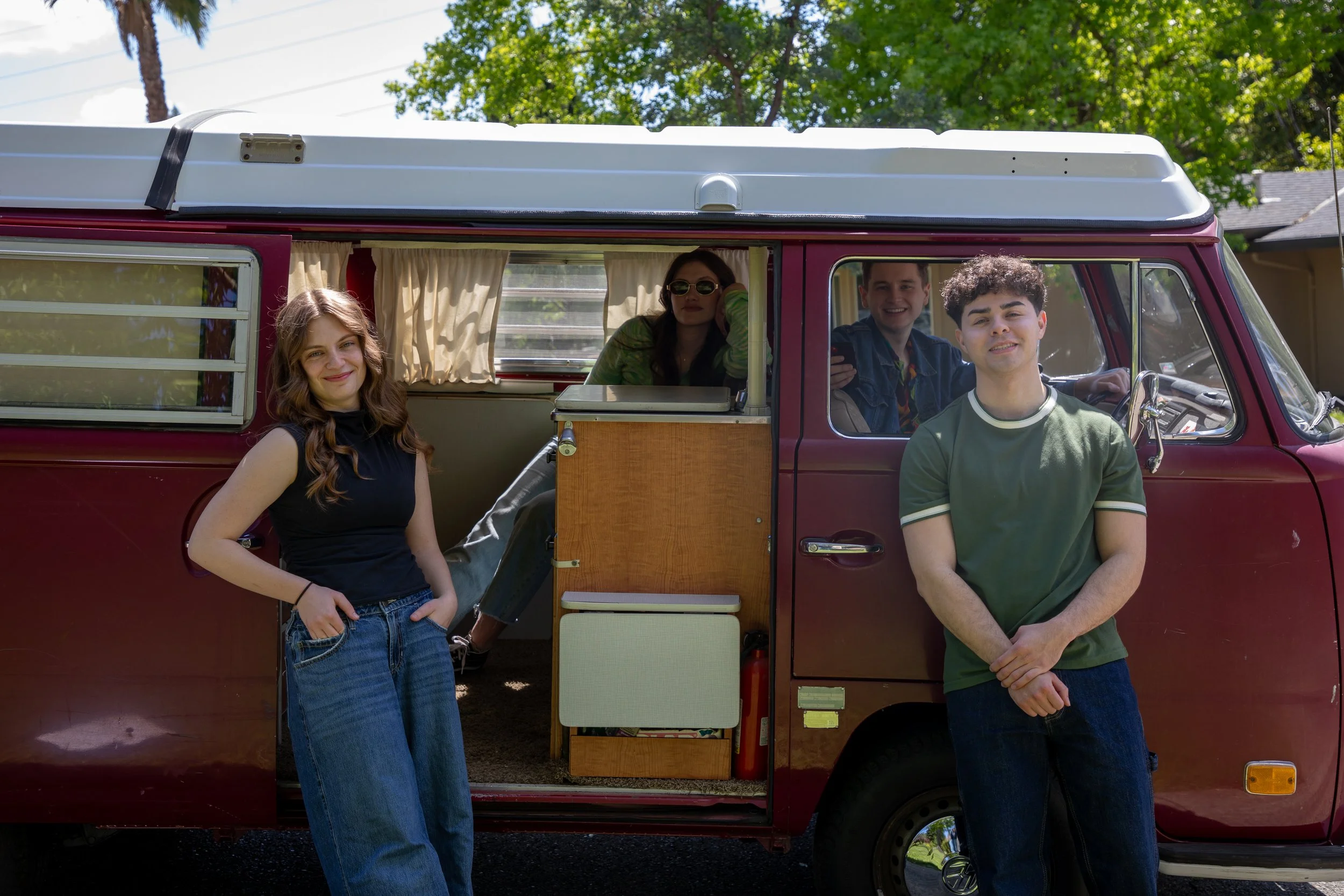Four young adults posing outside a vintage maroon camper van with a rooftop tent. Two are standing in front of the van, and two are inside near the windows, with a cozy curtain. They are smiling and enjoying a sunny day with green trees in the backgr