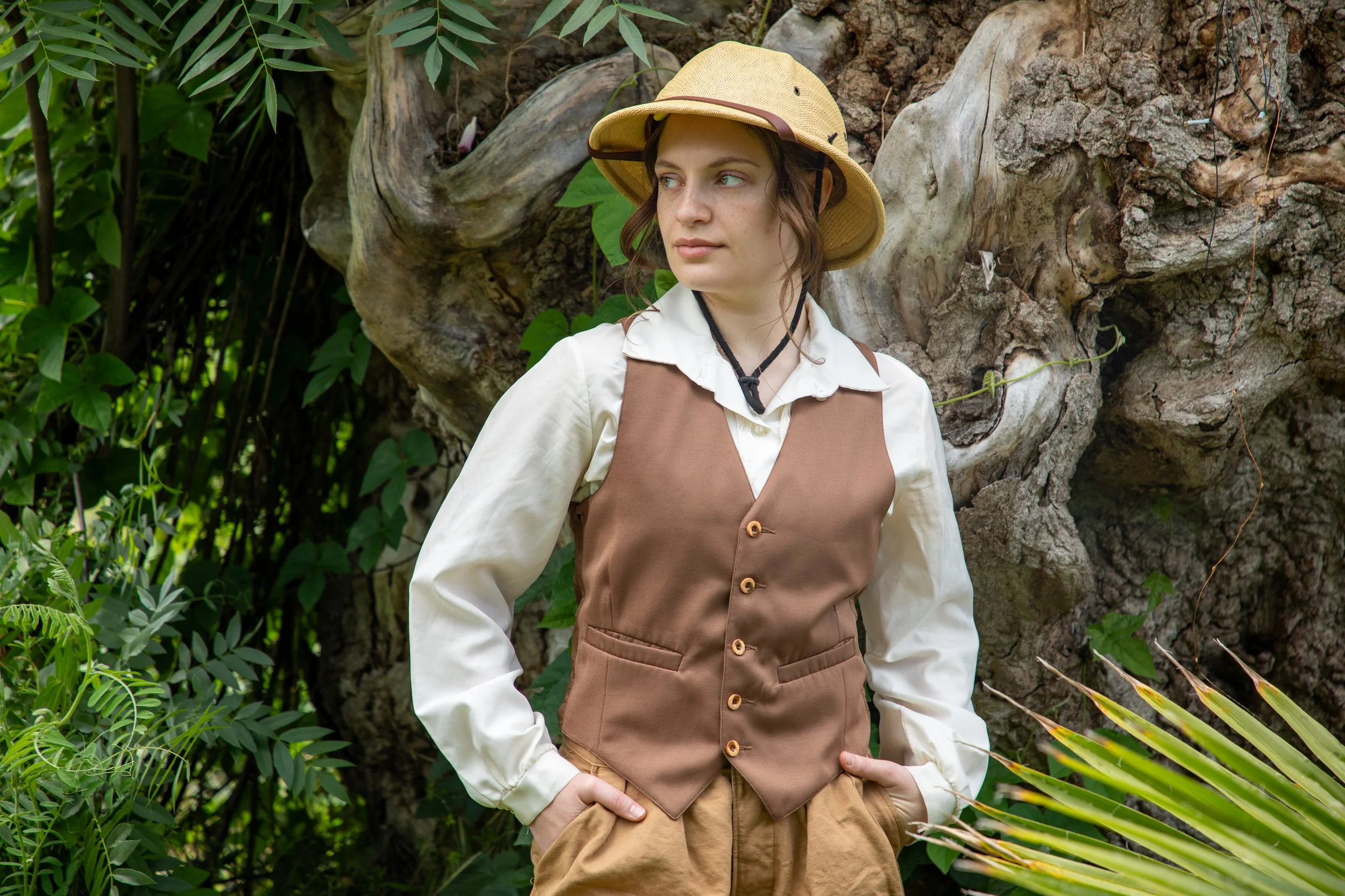 A young woman standing outdoors among green plants and trees, wearing a beige wide-brimmed hat, a white shirt, and a brown vest and pants, with her hands in her pockets is looking thoughtfully to the side.