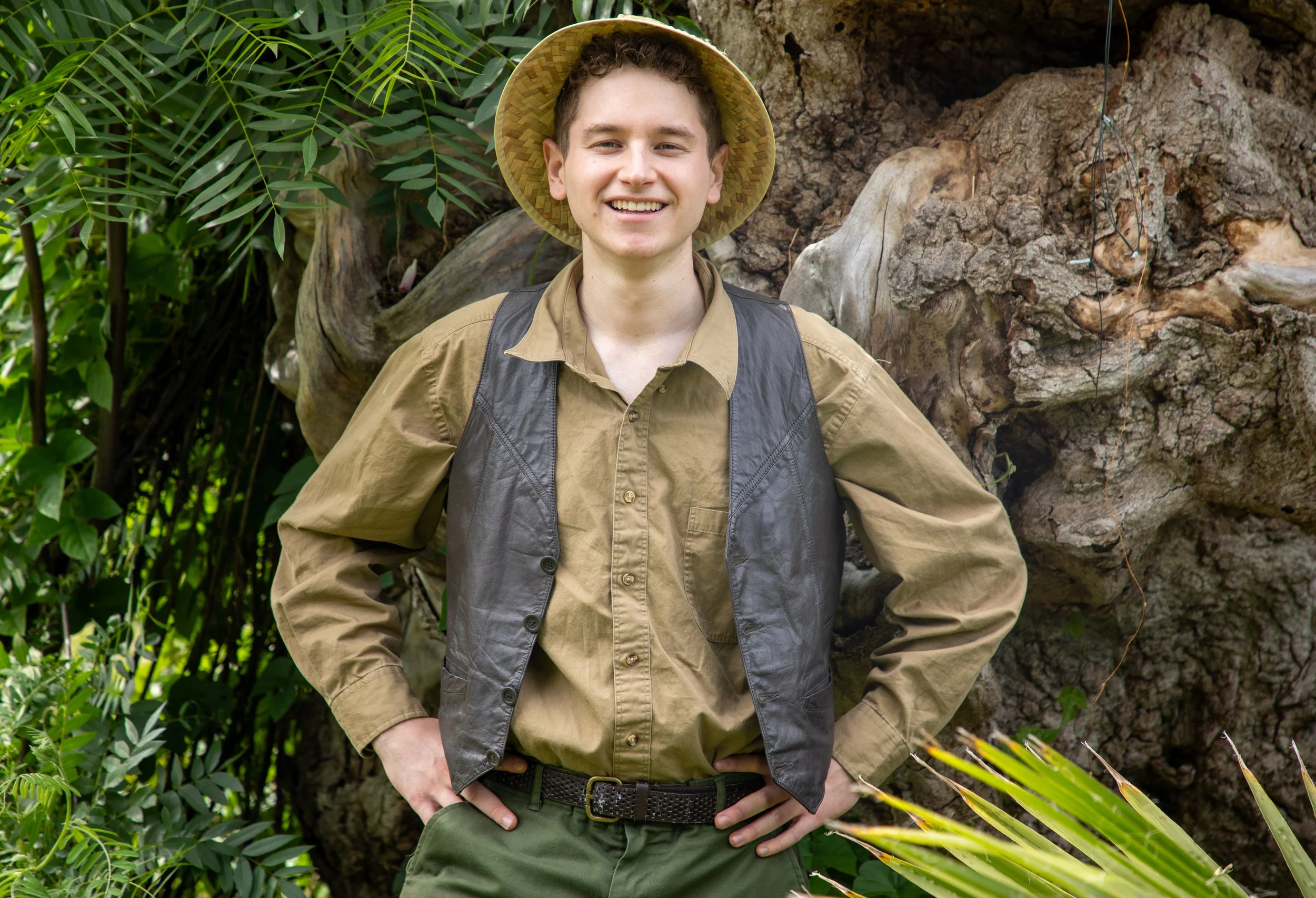 A young man with short curly hair, smiling, wearing a wide-brimmed straw hat, khaki shirt, and a black vest, standing outdoors among green plants and trees.