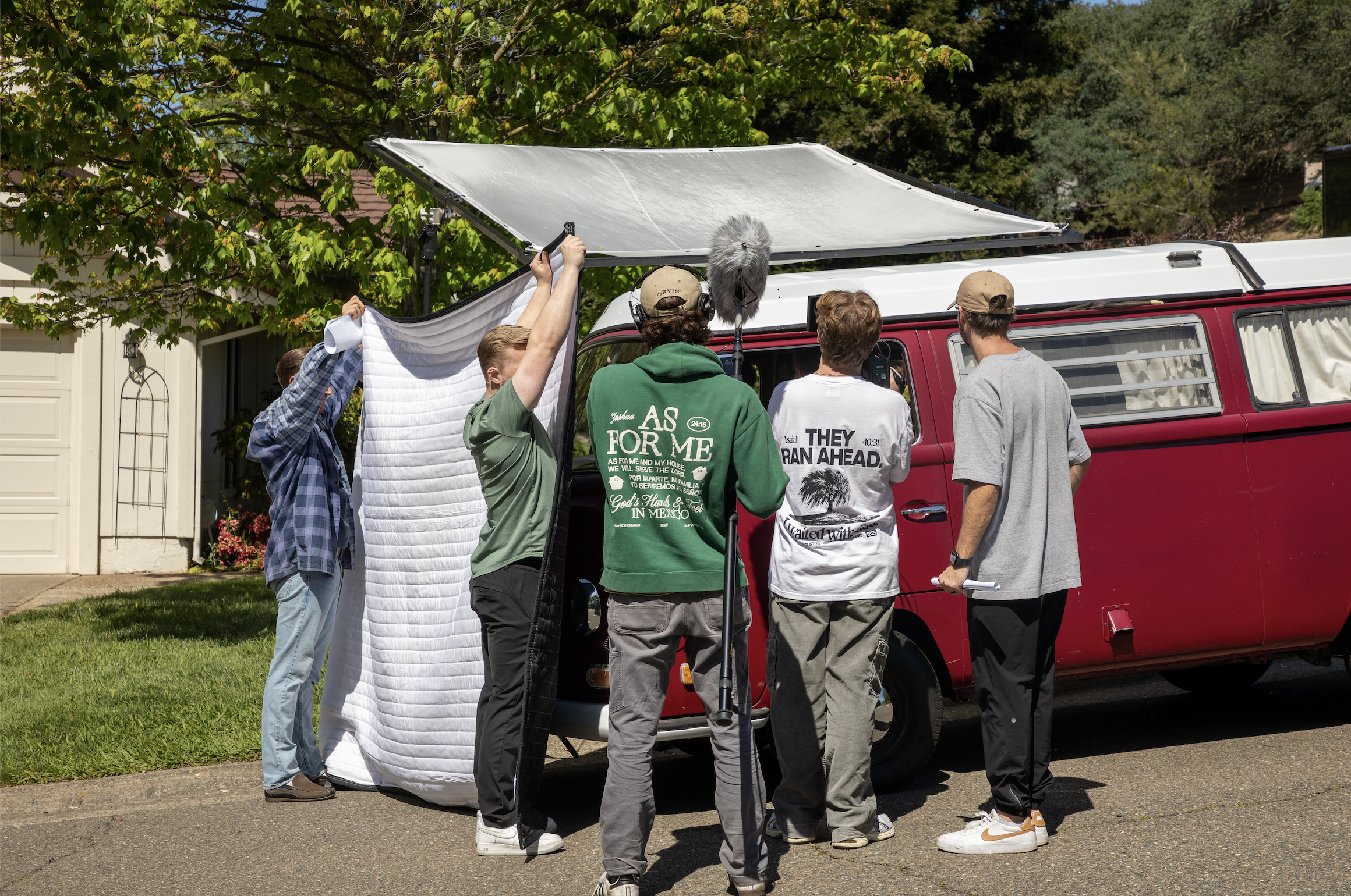 Film crew filming an outdoor scene next to a red van, with crew members setting up equipment and directing a large white sheet, in a residential neighborhood with trees and houses in the background.