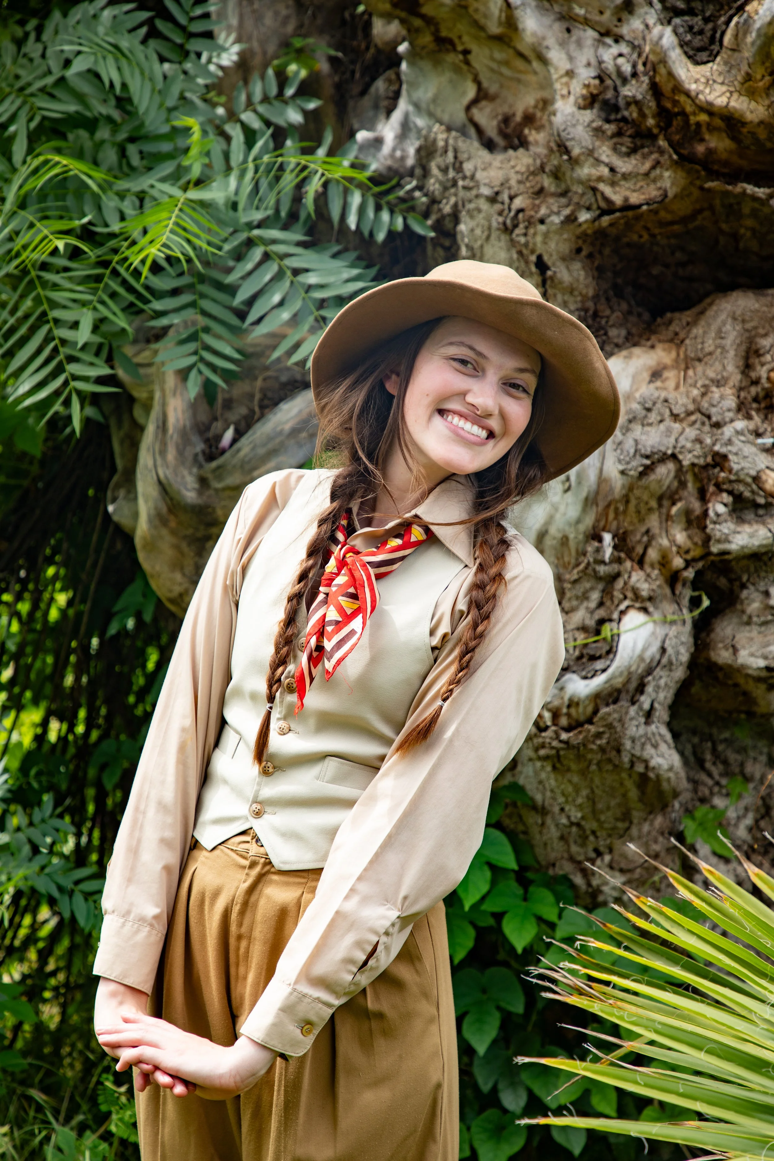 Young woman dressed as an explorer, wearing a beige uniform, a red and red neckerchief, and a wide-brimmed hat, standing in front of lush green foliage and a large tree trunk.