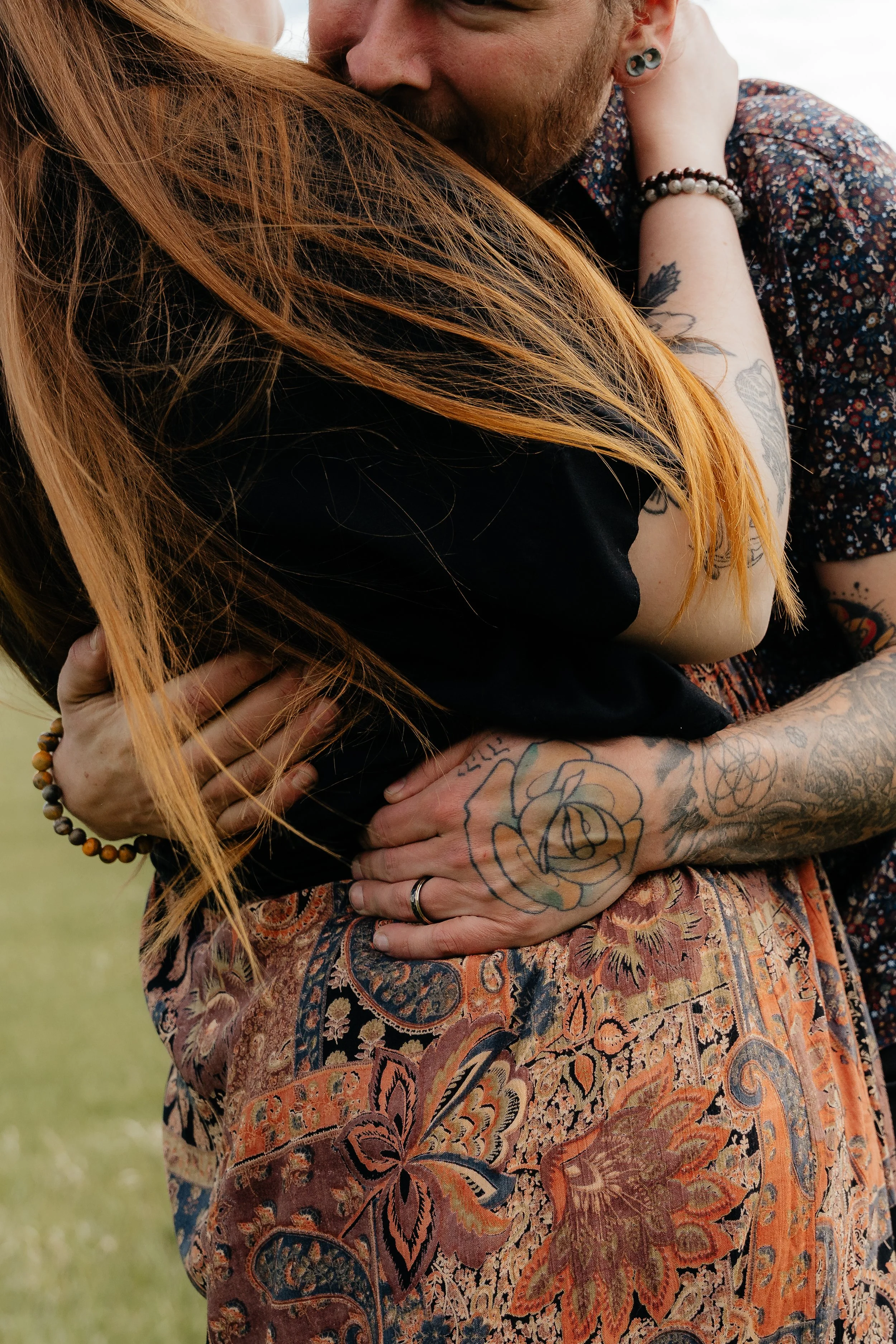 A couple hugging outdoors, with the woman’s long red hair and tattoos visible, and the man wearing a floral shirt and earrings.