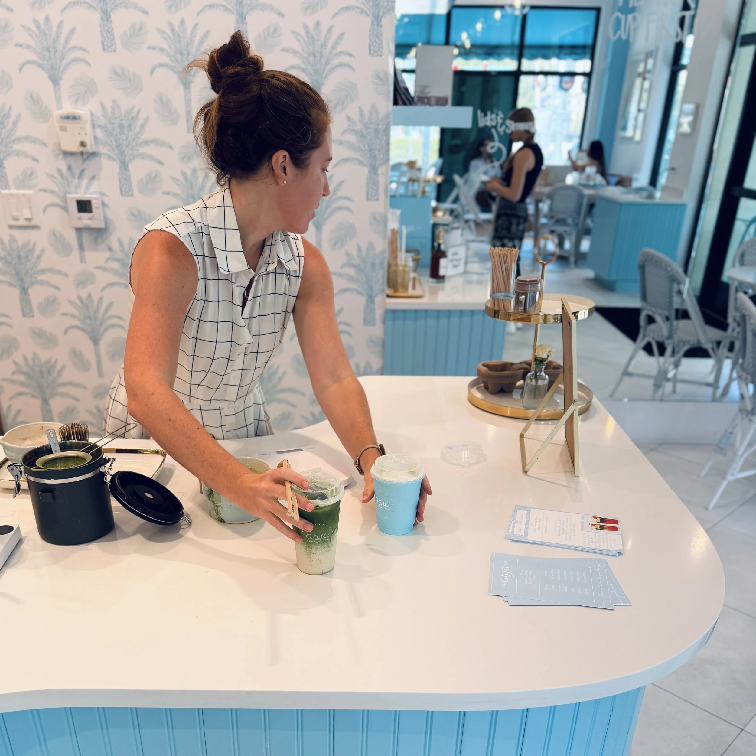 Sophie DeWaal stands behind Arya Tea Bar's curved pale blue counter, facing forward as she hands two drinks across. Nothing blocks the view of the workspace behind her.