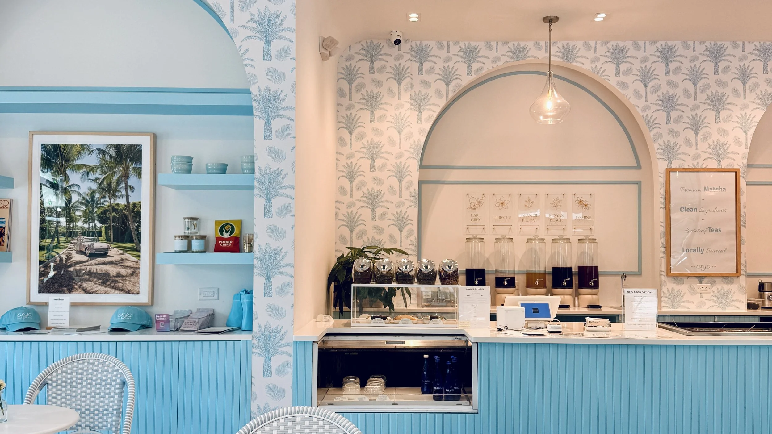 The interior of Arya Tea Bar. Pale blue palm-print wallpaper, an arched alcove with blue shelving, white bistro tables and chairs, and glass pendant lights.