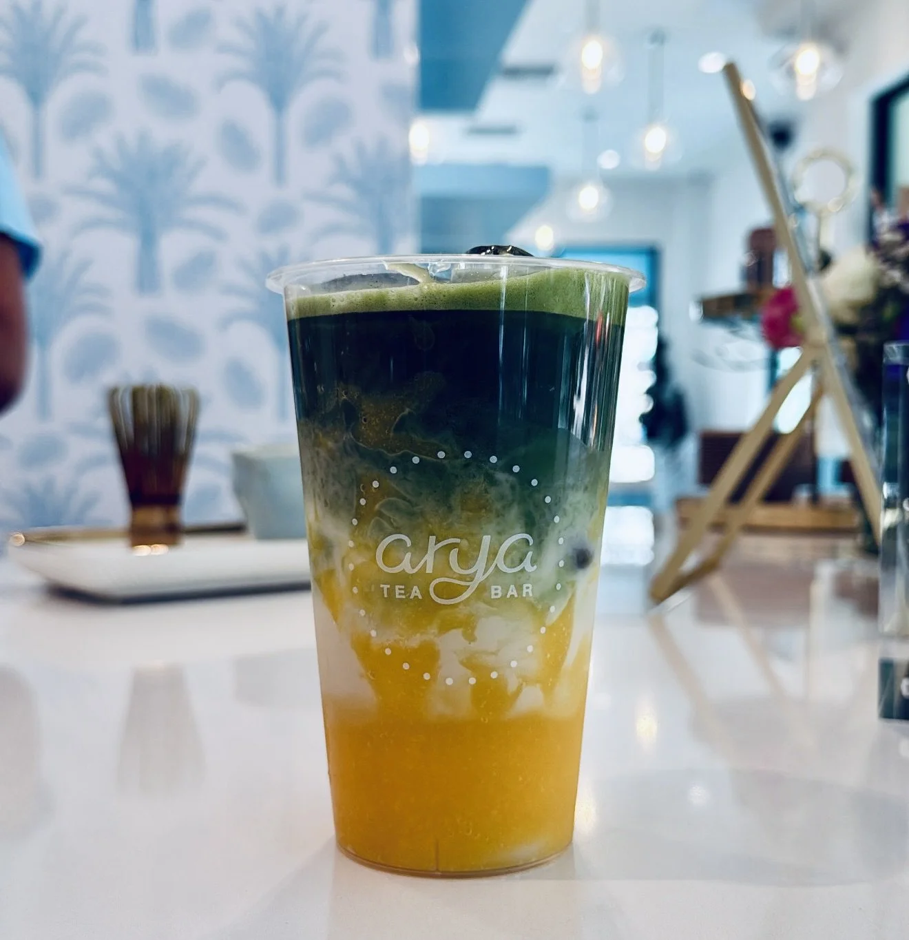 An iced mango matcha in an Arya Tea Bar cup printed with "the best summer I ever had was a winter in Palm Beach," beside fresh white daisies in a small glass vase tied with a blue ribbon.