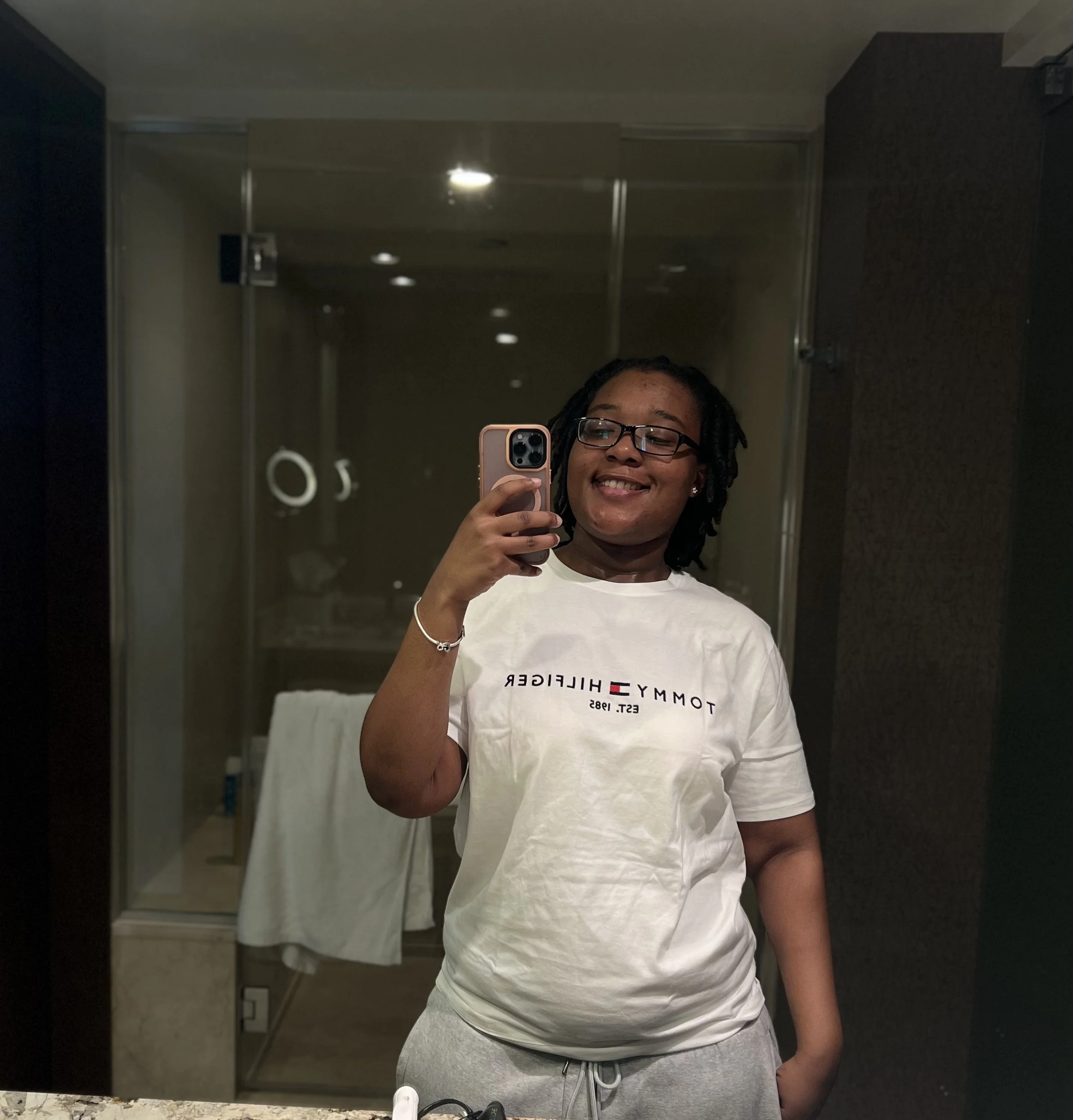Young person with glasses taking a mirror selfie in a bathroom, wearing a white Tommy Hilfiger T-shirt and gray sweatpants.