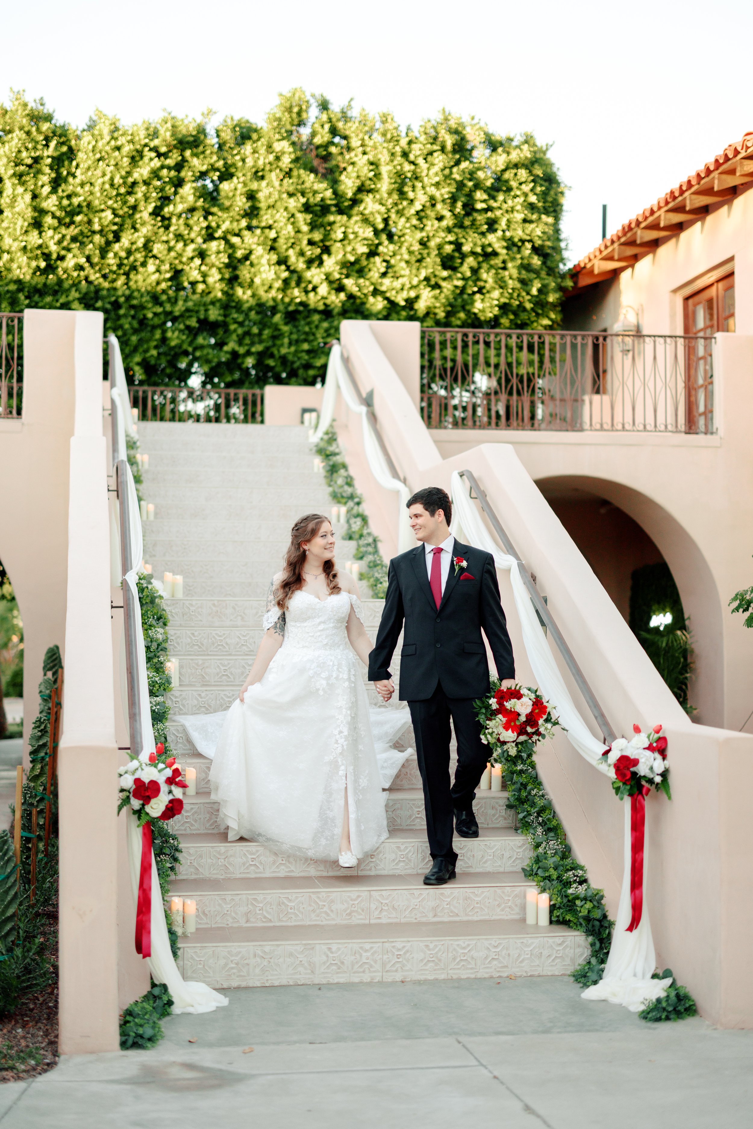 A bride and groom walking down decorated outdoor stairs holding hands, with candles and floral arrangements, in a wedding setting.
