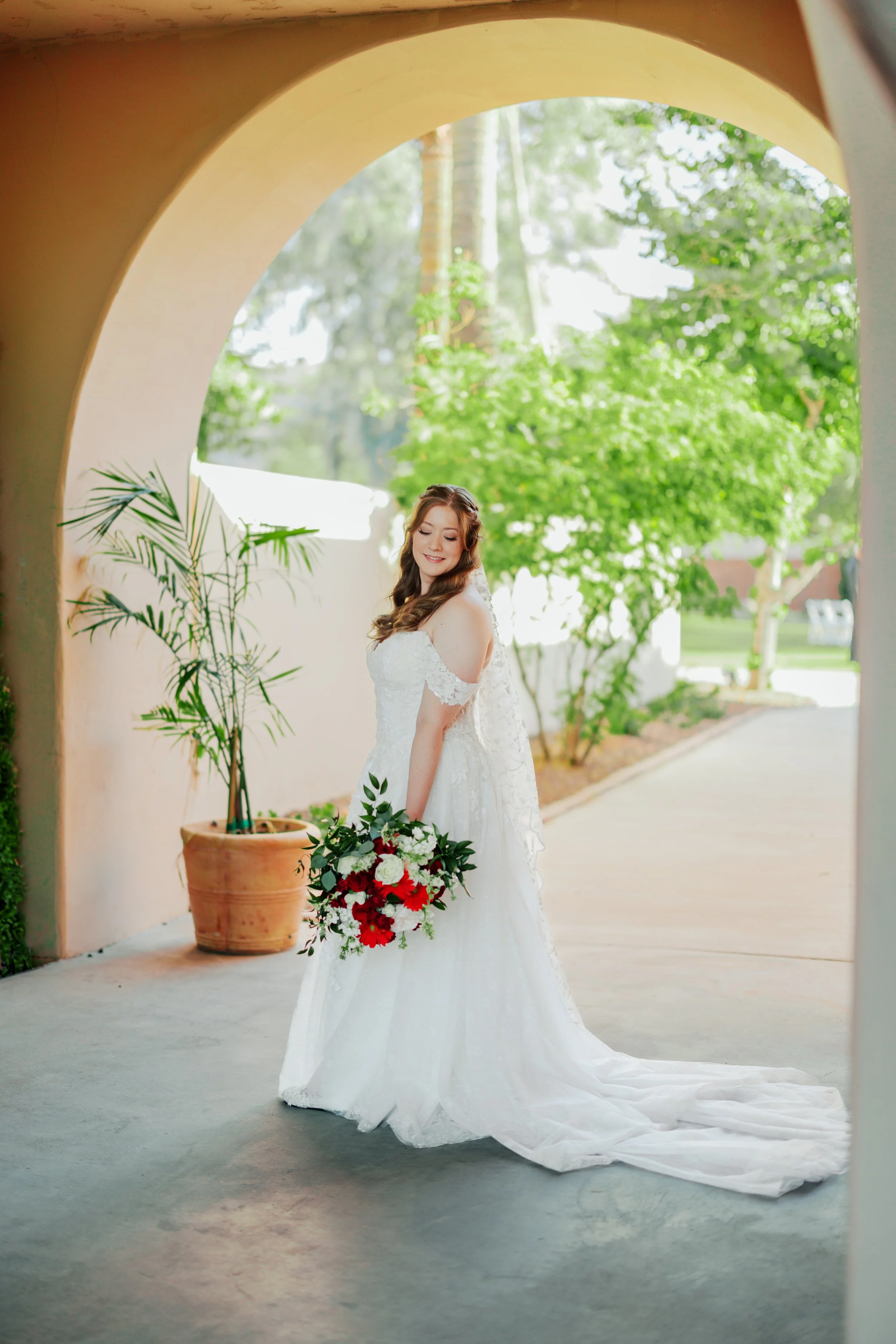 A bride in a white wedding dress holding a bouquet of red and white flowers, standing under an archway with a background of green trees and sunlight.