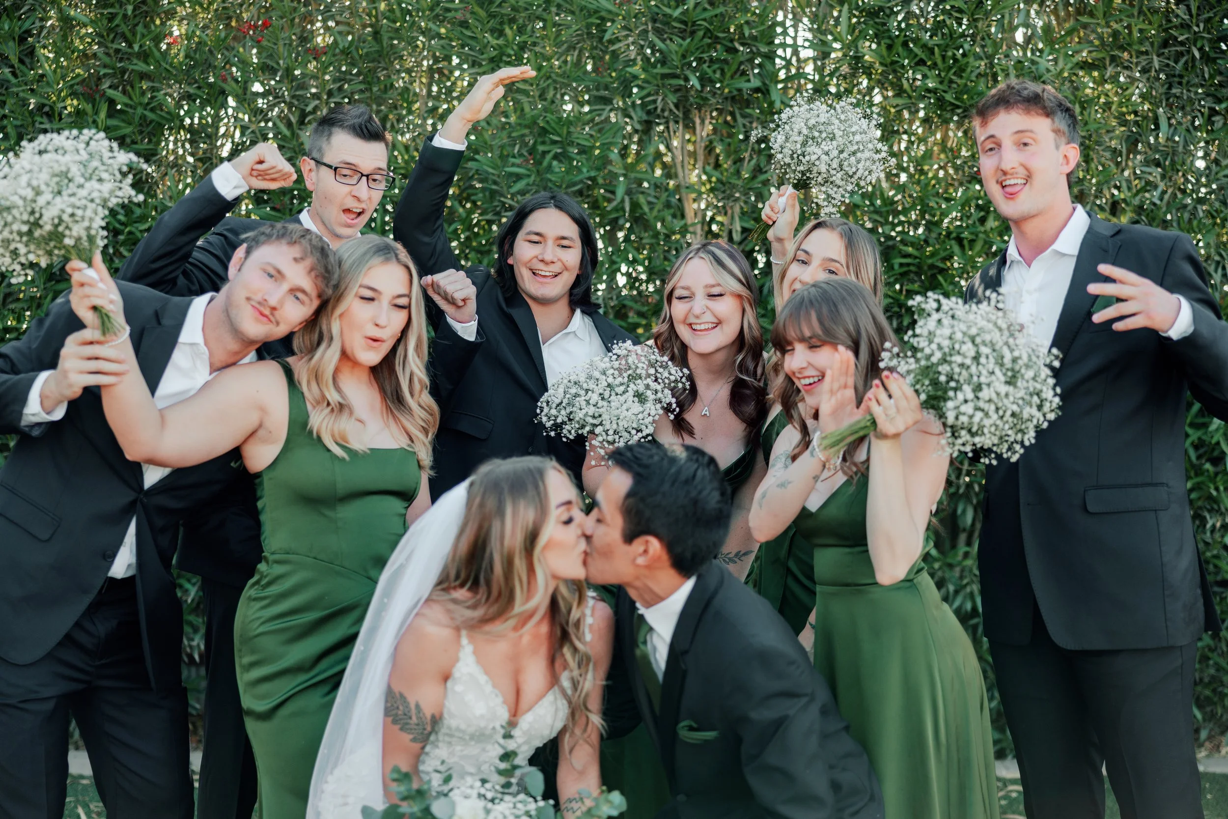 A bride and groom kiss surrounded by friends at a wedding celebration; the friends are smiling, cheering, and holding bouquets of white flowers.