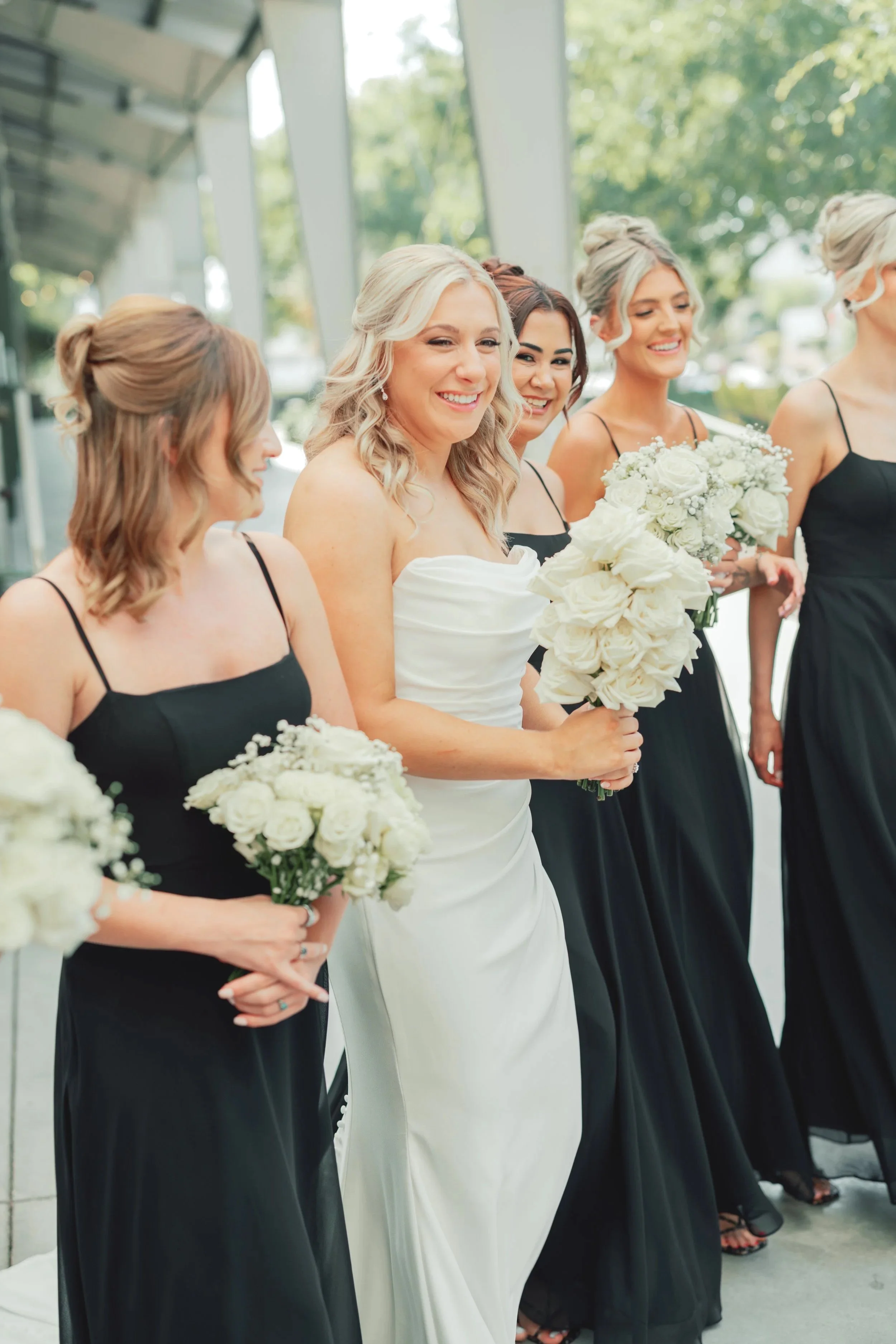 Bride in white wedding gown holding a bouquet of white roses, surrounded by bridesmaids in black dresses holding similar bouquets, outdoors on a sunny day.