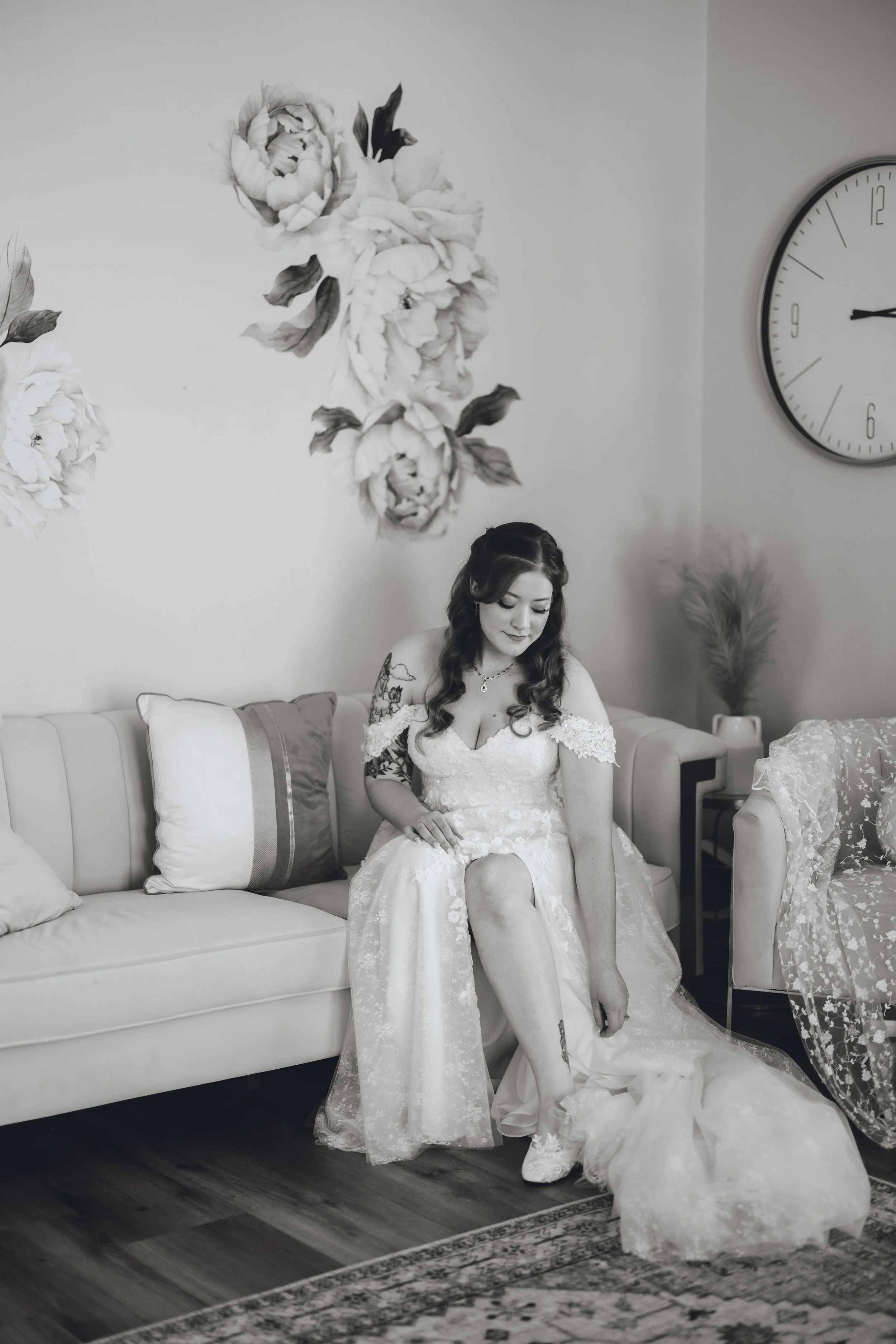 A woman in a wedding dress sits on a sofa, adjusting her shoe, with a clock on the wall and floral wall art behind her.