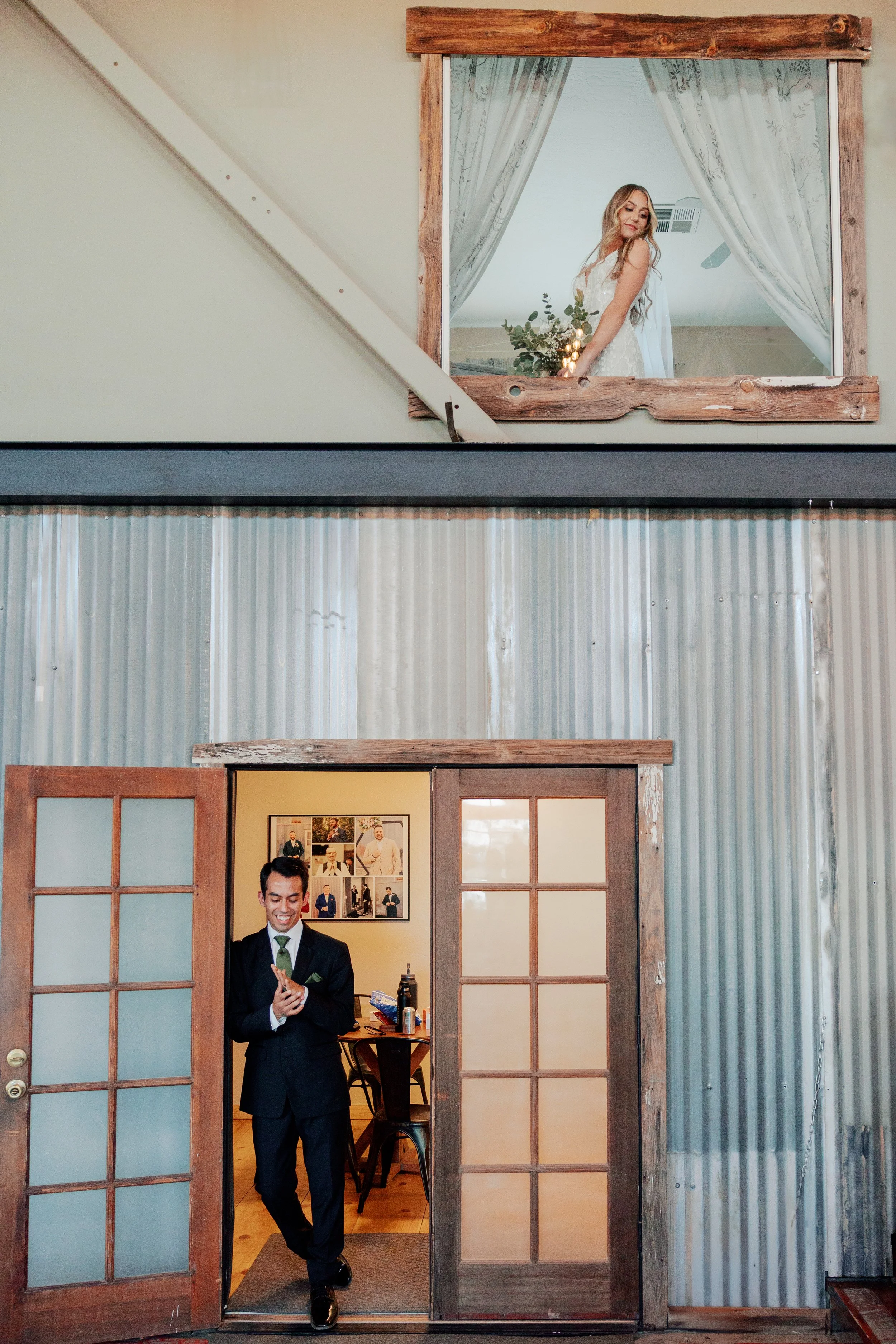 A bride looking out of a rustic wooden-framed window above a sliding door, holding a bouquet, smiling. A groom in a dark suit standing in a room with corrugated metal walls, smiling, with photos on the wall behind him.