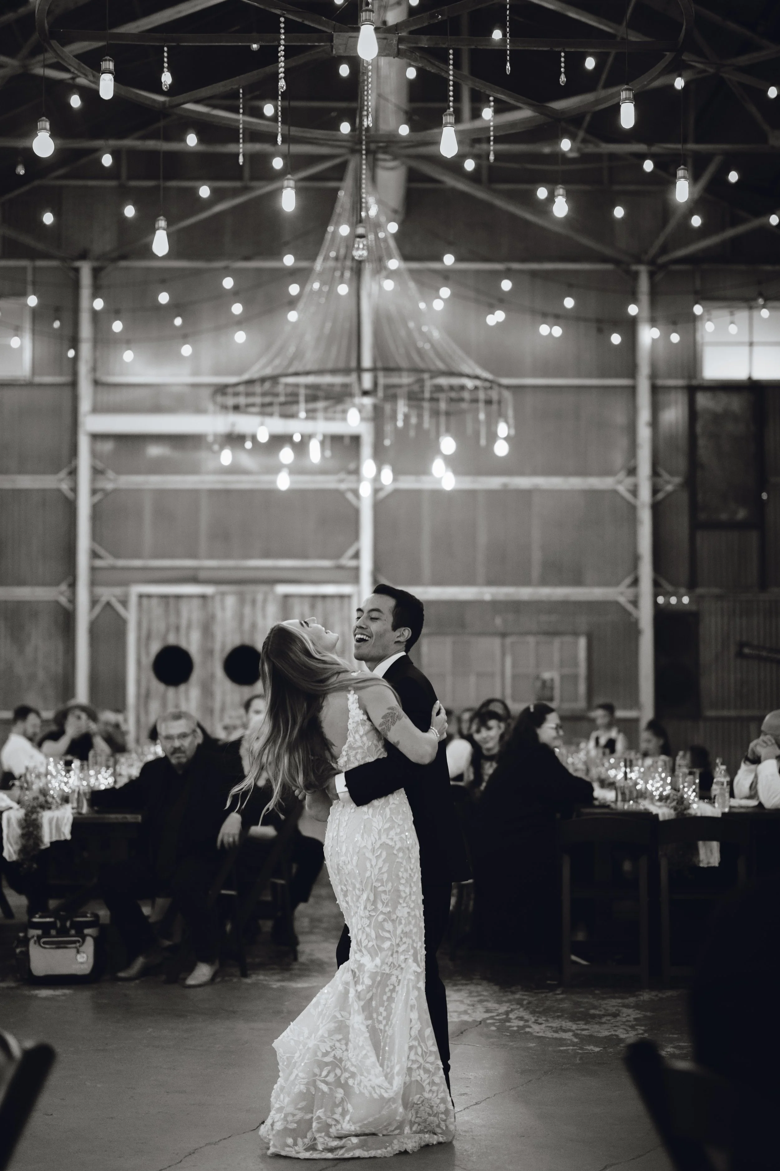 A bride and groom dancing at their wedding reception in a rustic barn decorated with string lights, with guests seated at tables watching in the background.