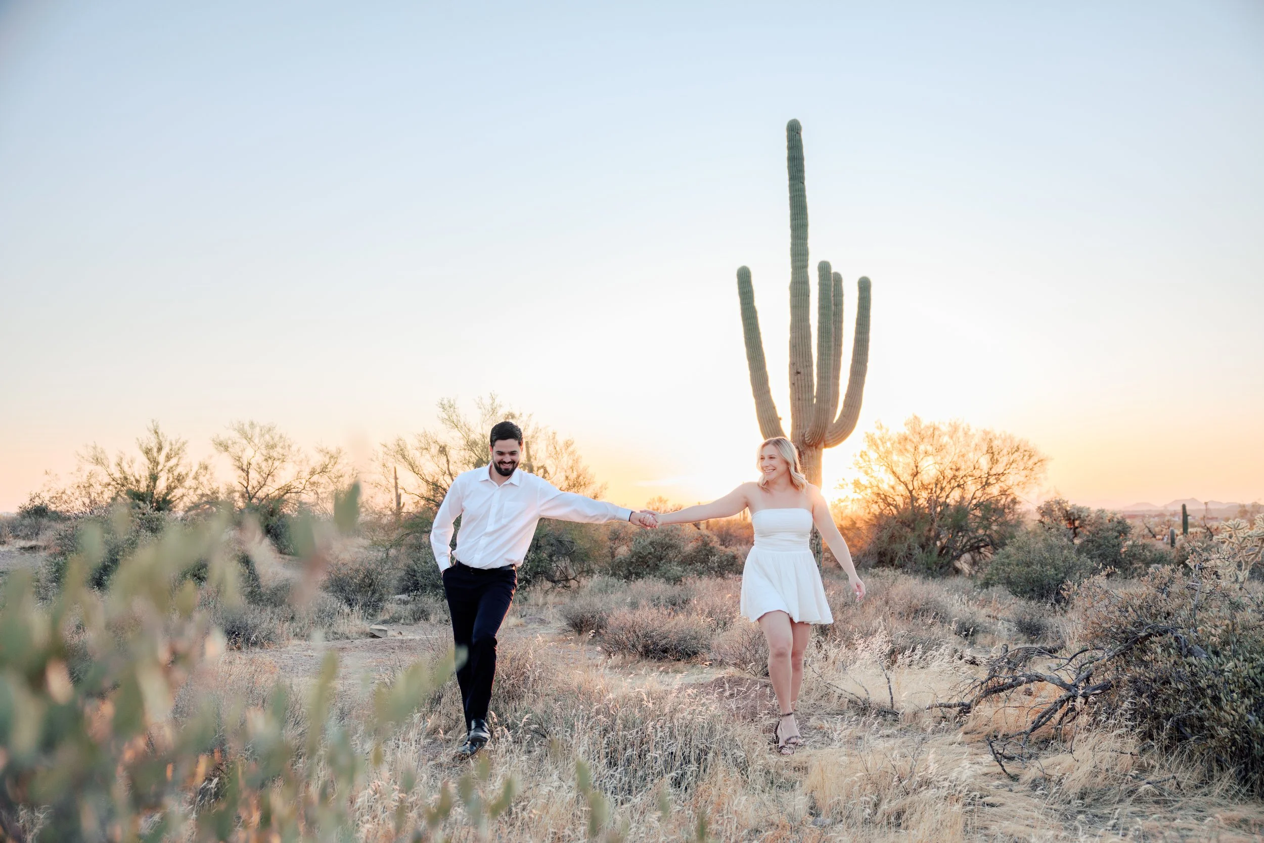 A smiling couple holding hands and walking in a desert landscape at sunset, with a large saguaro cactus in the background and dry bushes and grass surrounding them.