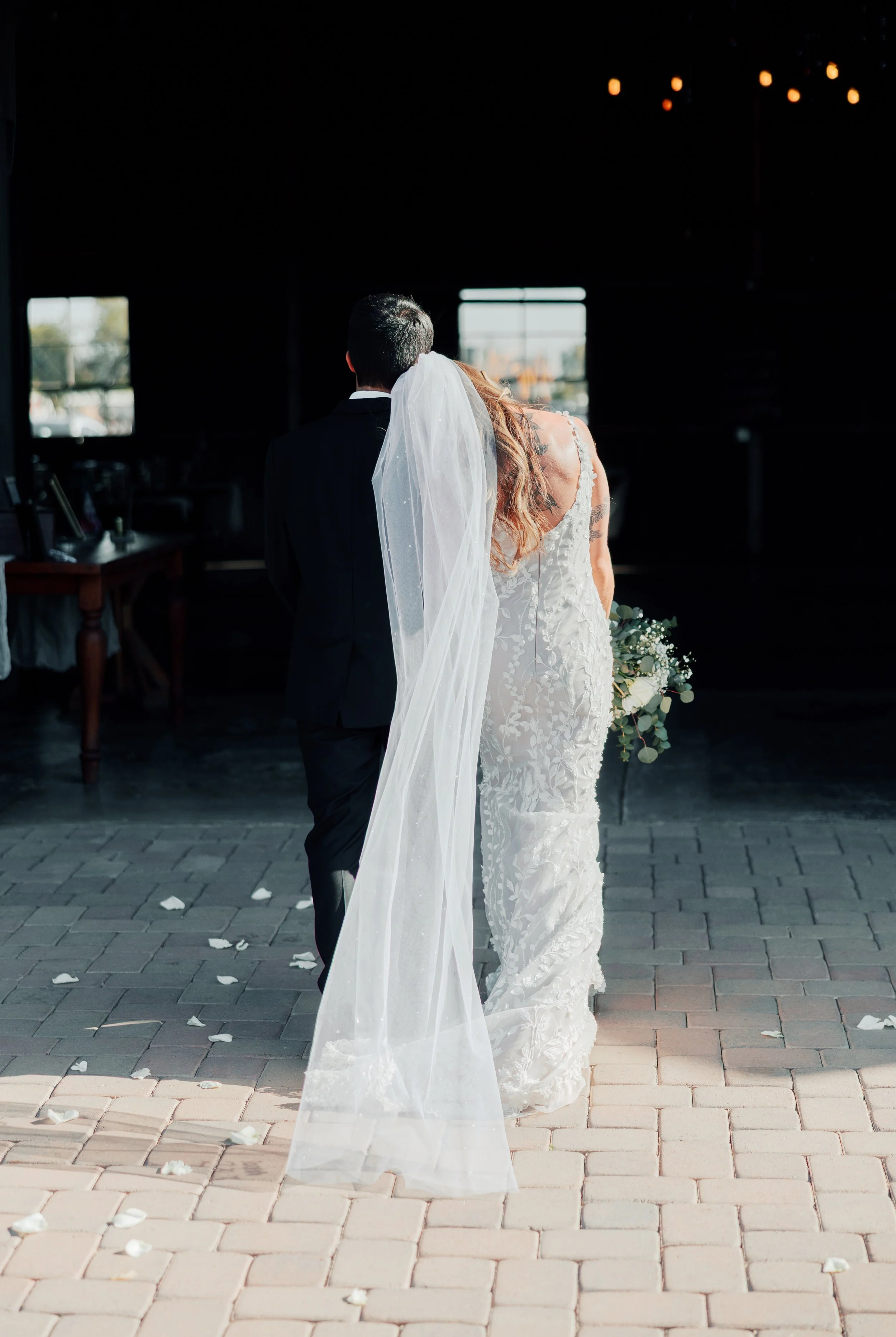 Bride and groom walking together after their wedding ceremony, bride holding a bouquet, inside a venue with brick floor and open doors, petals scattered on the ground.