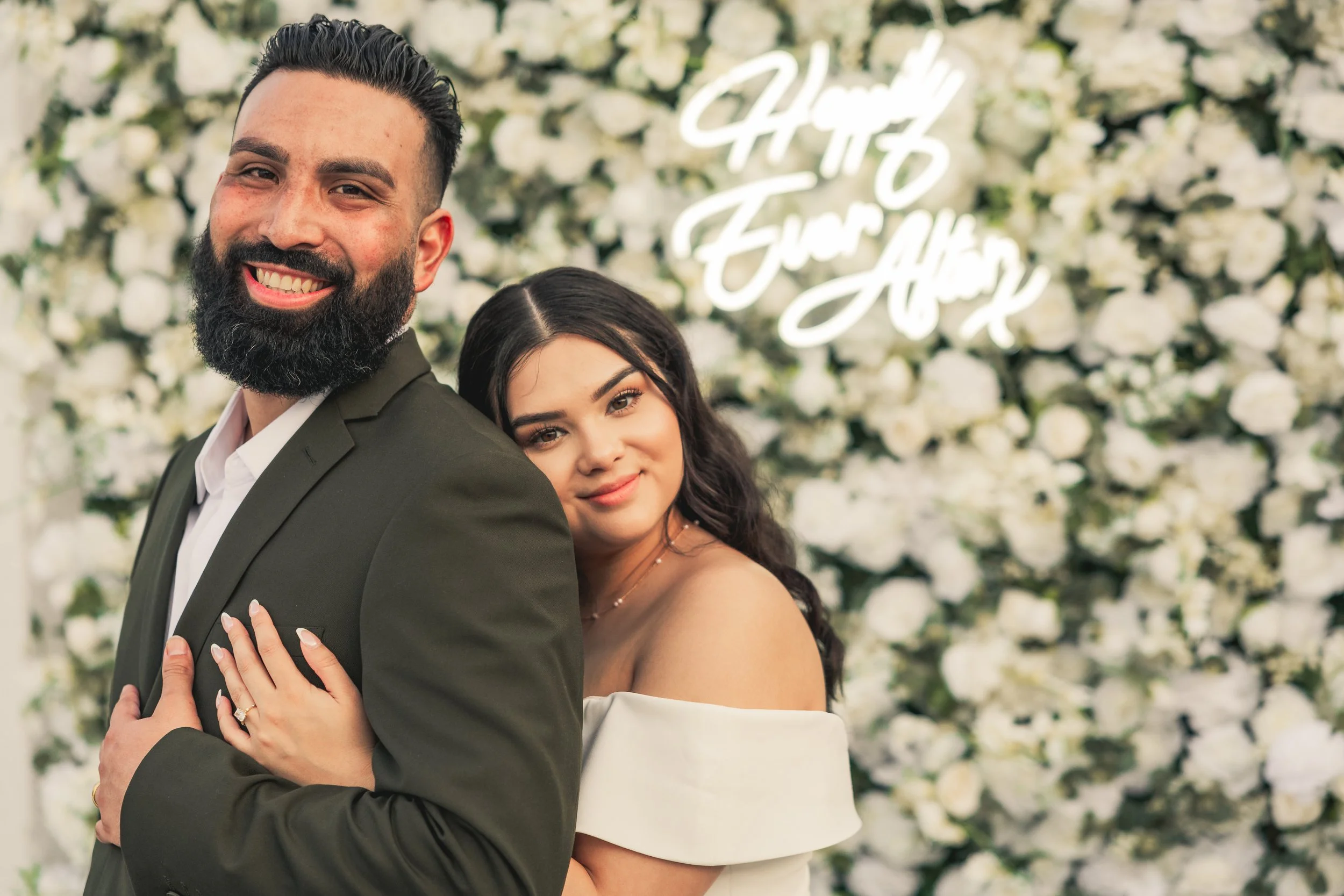 A smiling man with a beard in a suit and a woman with dark hair in an off-shoulder white dress, standing close together at a wedding with a white floral backdrop and a neon sign that says 'Happy Ever After'.