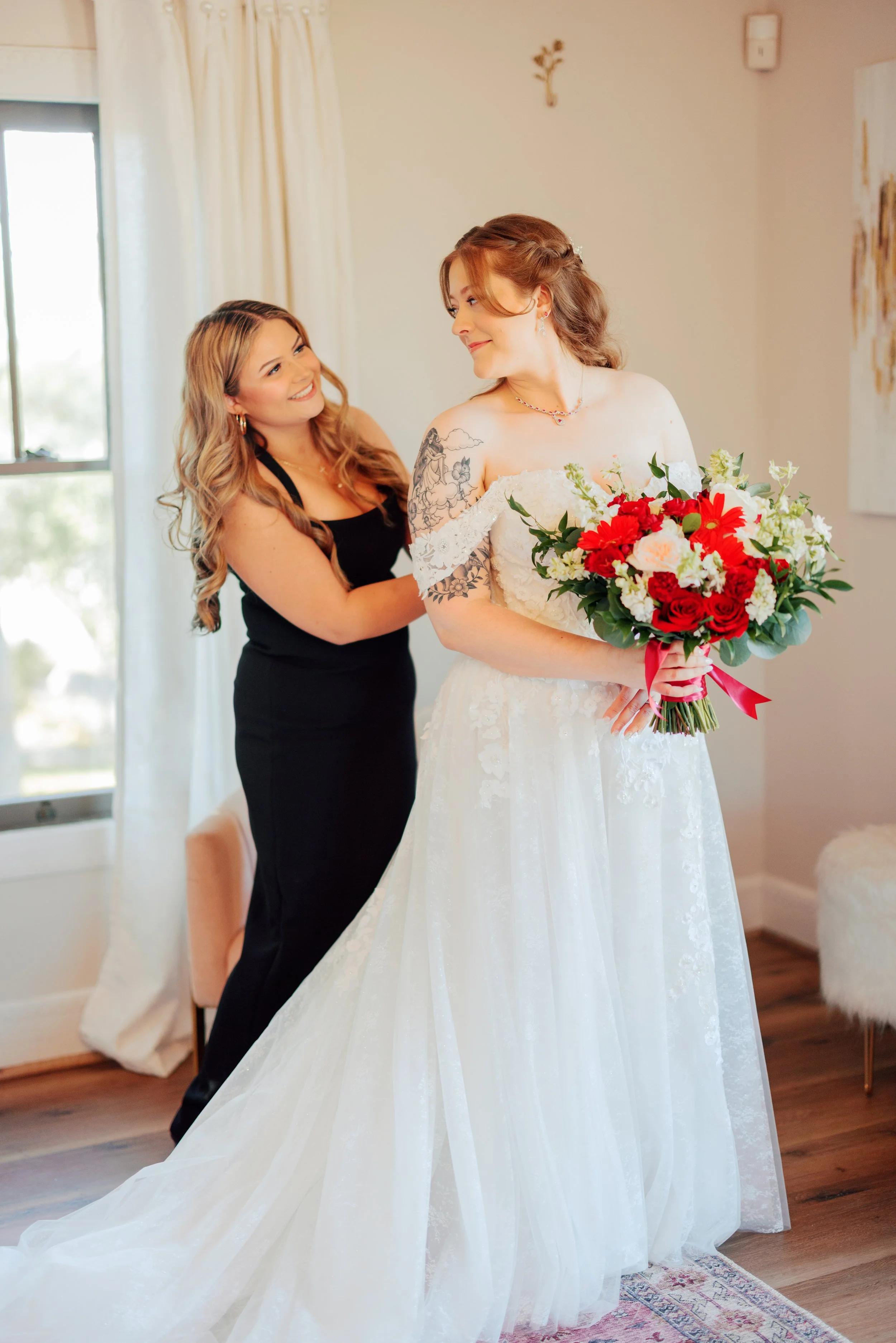A bride in a white wedding dress holding a bouquet of red, white, and pink flowers, standing in a room with a woman in black helping her adjust her dress.