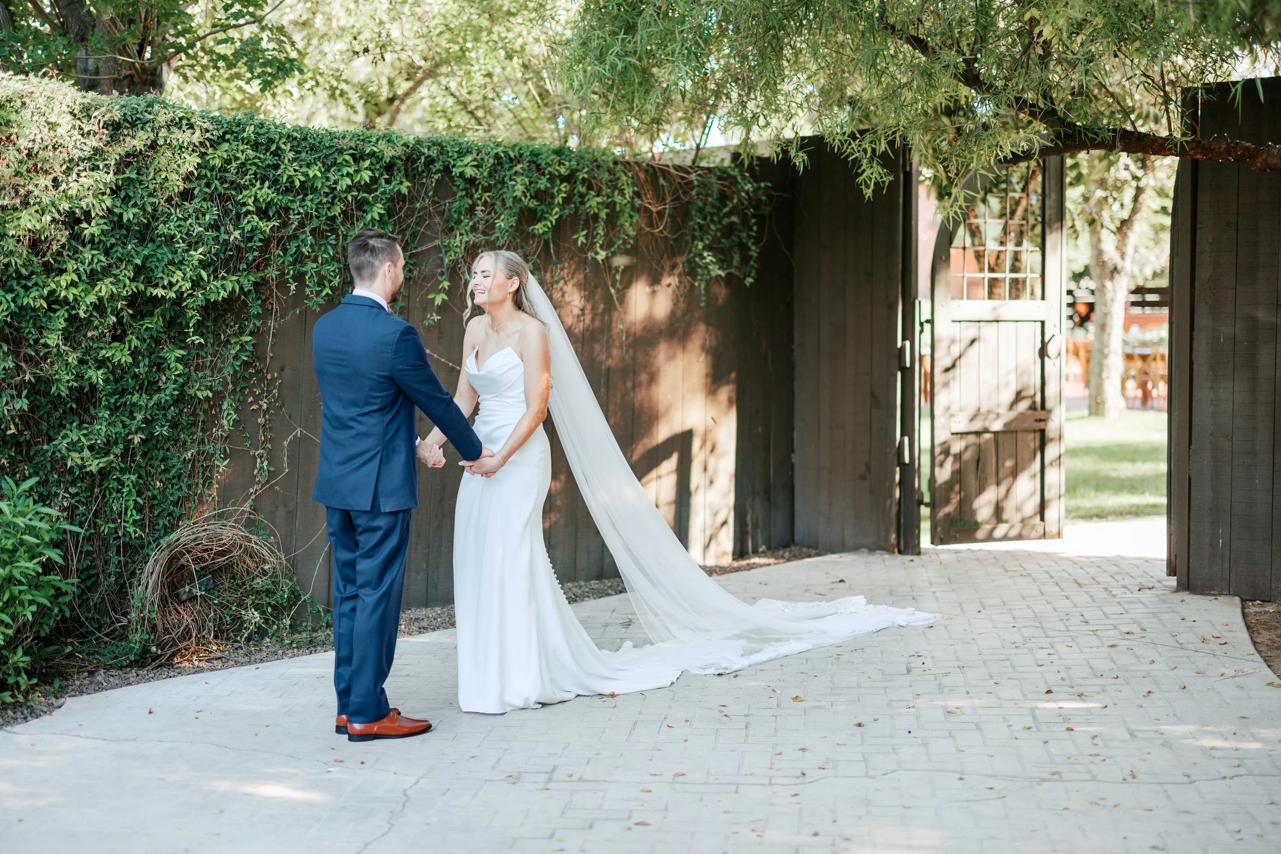 A bride and groom holding hands and smiling at each other outdoors at their wedding, standing on a paved path with a wooden fence and open gate in the background, surrounded by trees and greenery.