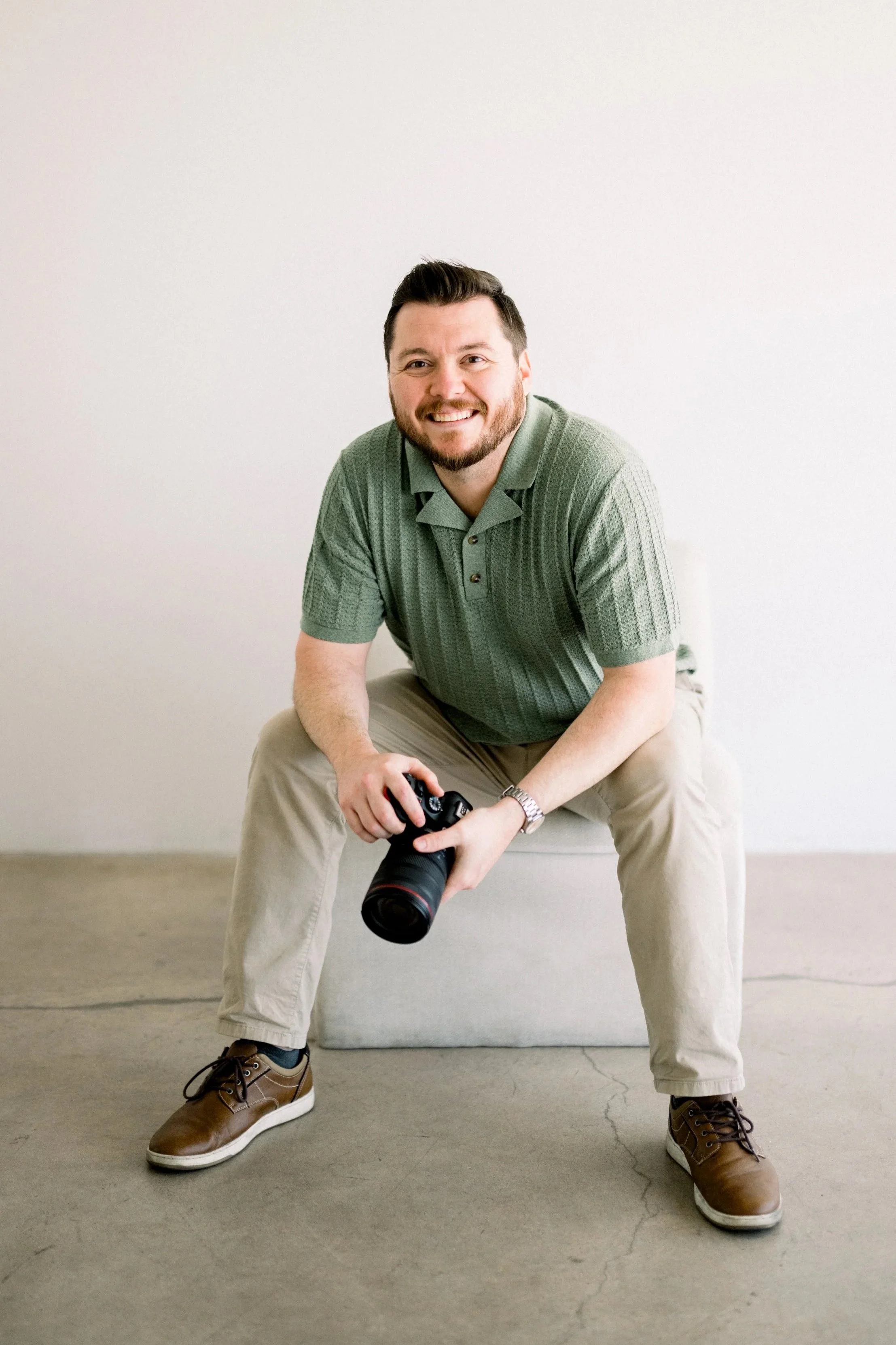 A man with a beard and short dark hair, wearing a green polo shirt and beige pants, sitting on a small gray stool and holding a camera in his right hand, smiling at the camera against a plain white wall.
