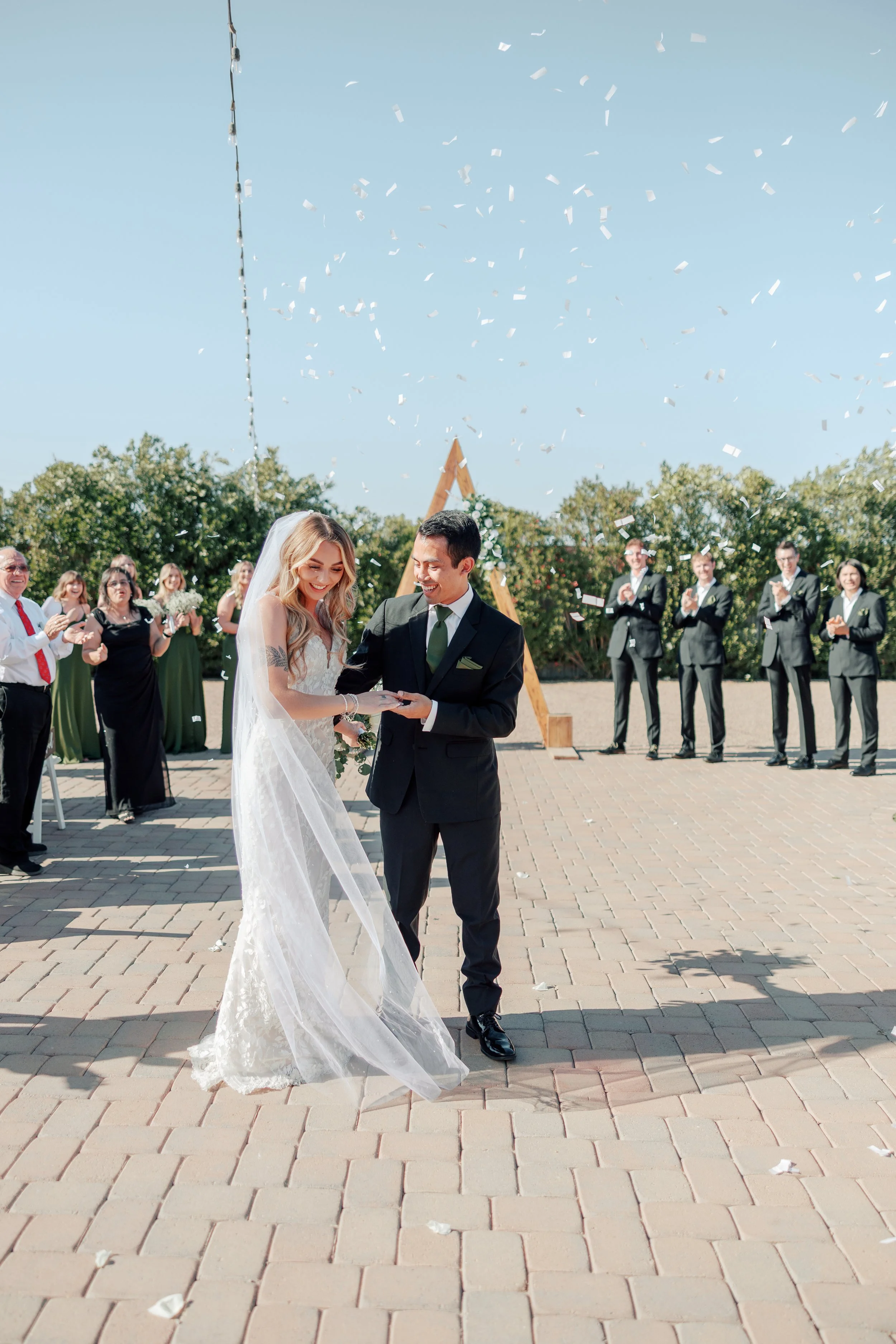 Bride and groom holding hands and walking down the aisle during an outdoor wedding ceremony as guests celebrate beneath a blue sky with falling confetti.