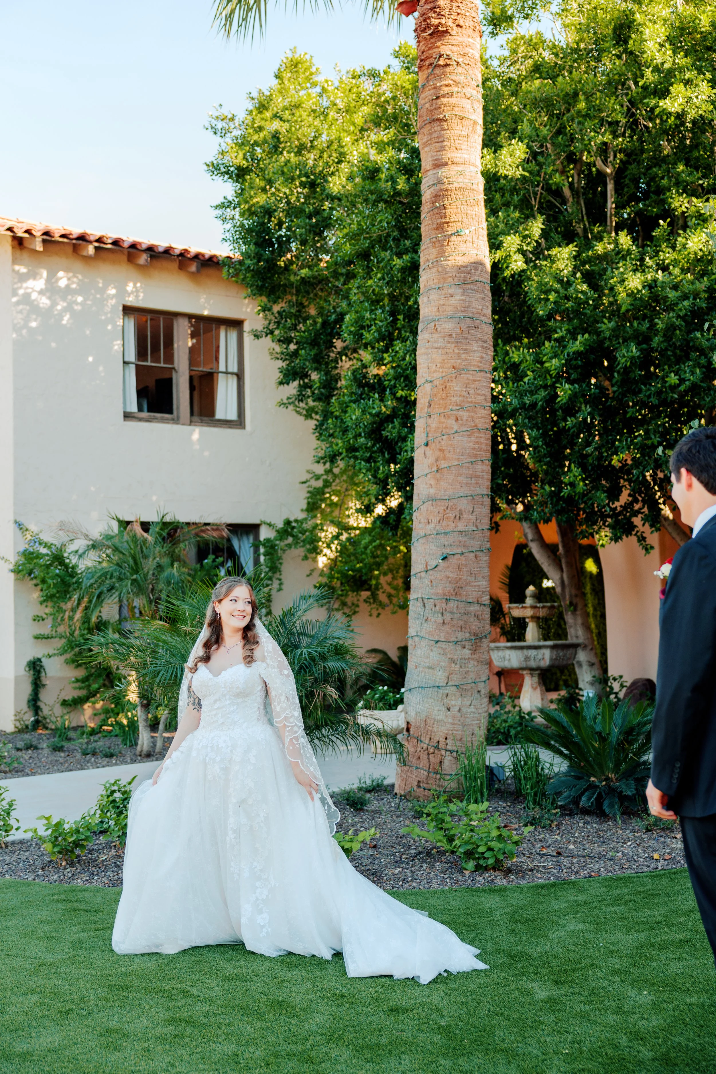 Bride in a white wedding dress standing outside, smiling at her groom who is wearing a black suit, against a backdrop of green trees and a white building.