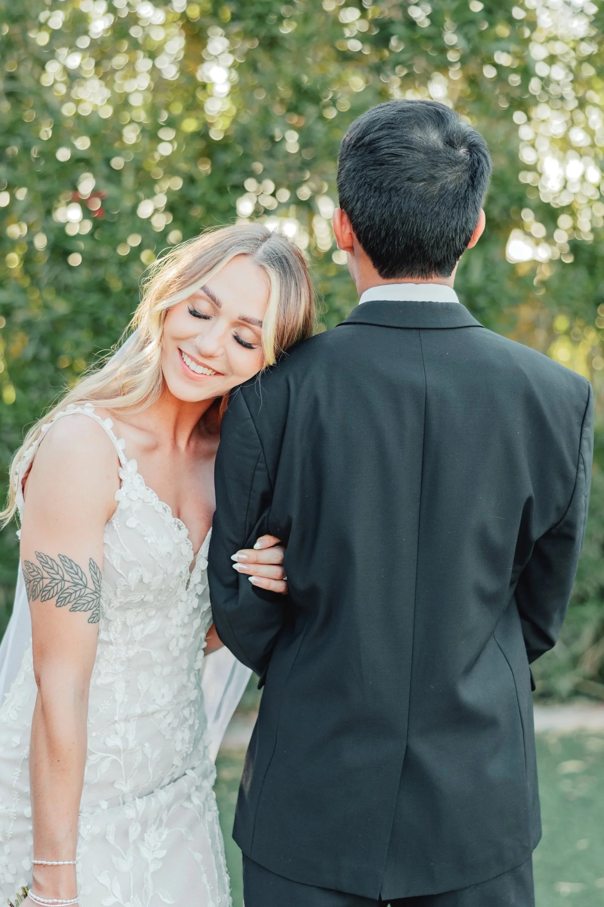 A happy bride with long, wavy blonde hair and a white wedding dress, smiling with her eyes closed, holding onto a groom in a black suit, outdoors with green trees and dappled sunlight in the background.