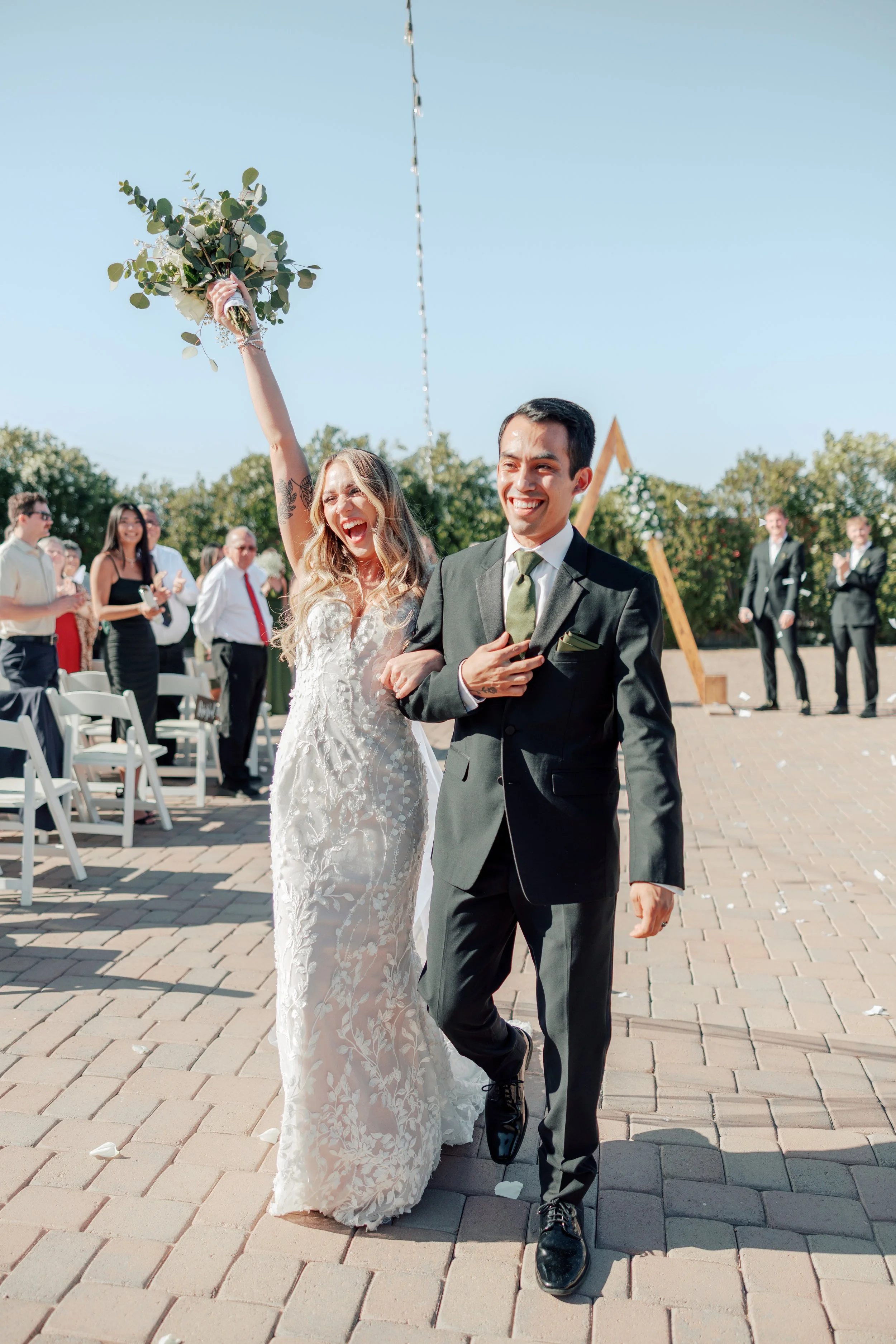 A newlywed couple, the bride in a white lace dress and the groom in a black suit, happily walking arm in arm after their wedding ceremony outdoors on a sunny day, surrounded by friends and family.