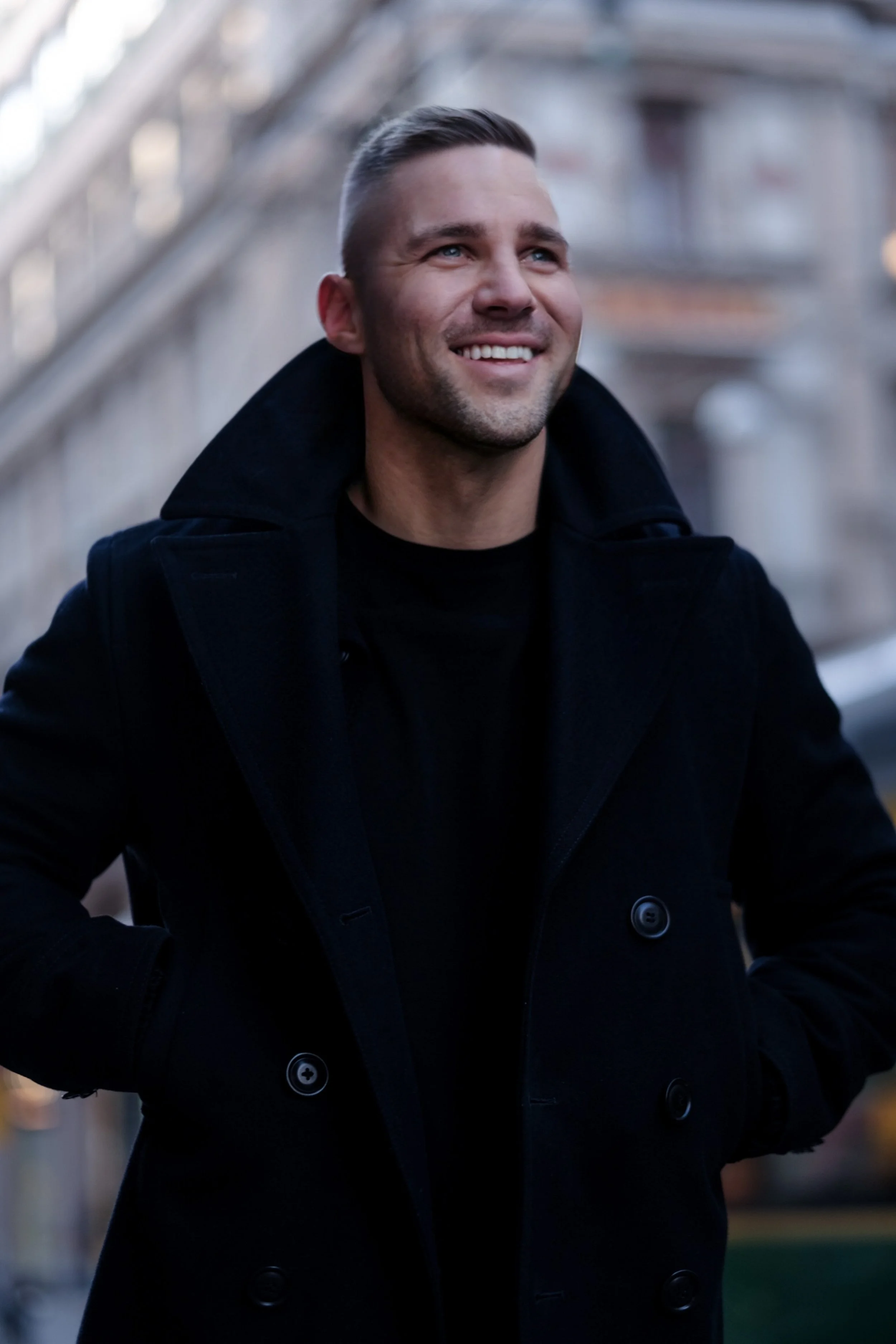 A smiling man with short hair wearing a black coat outdoors.