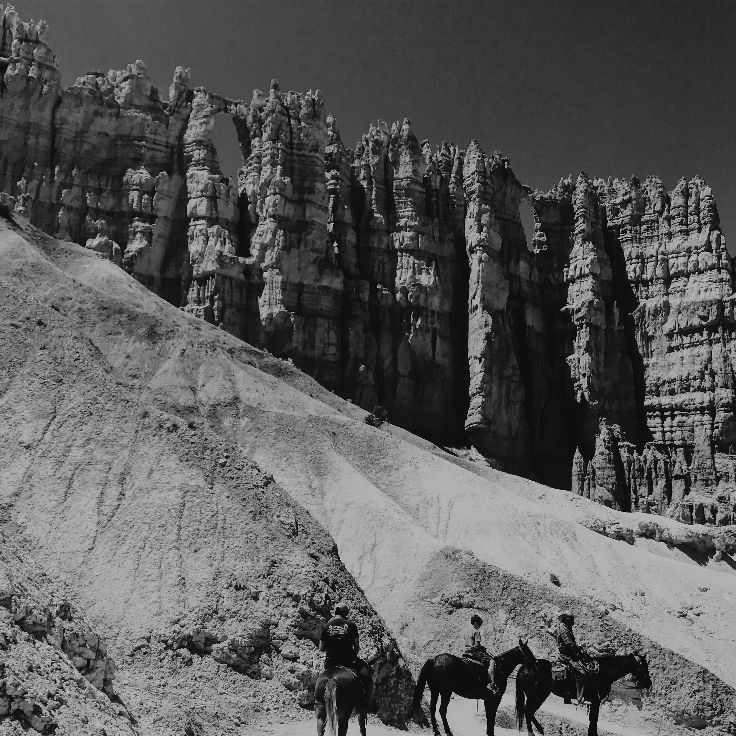 Three riders on horses in front of tall, rugged rock formations in a desert landscape.