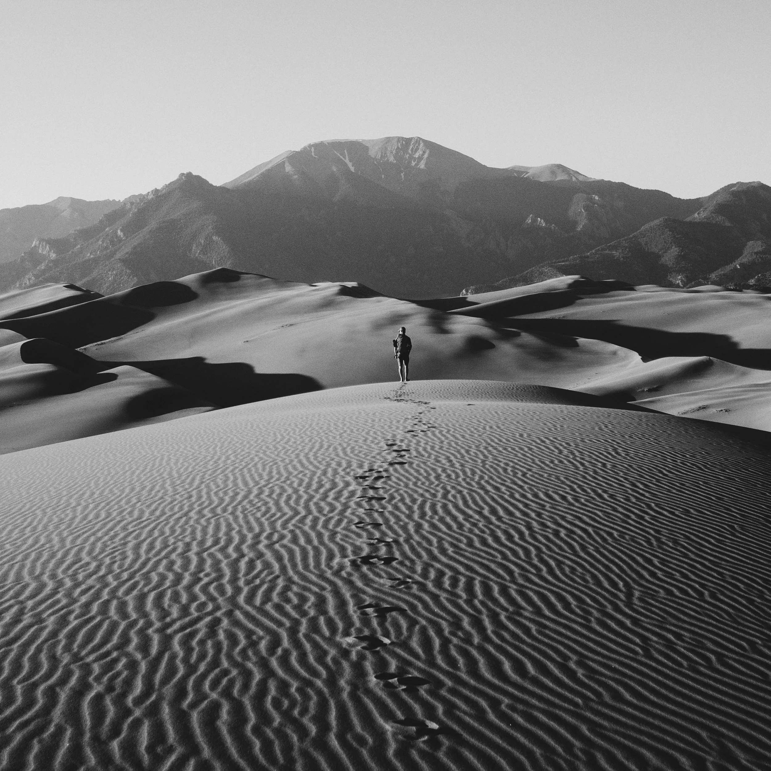 Person walking on sand dunes with mountains in the background, black and white.