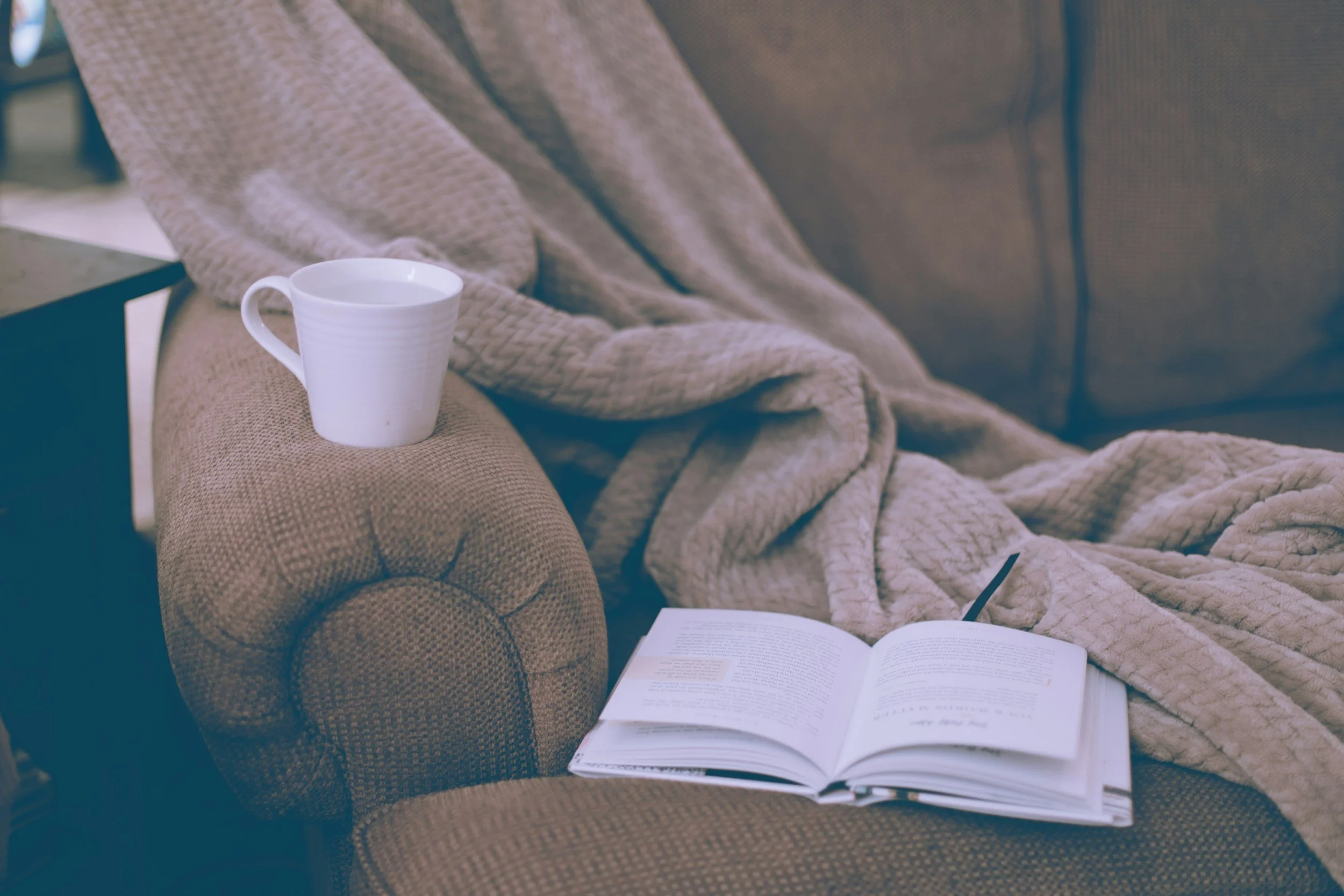 A cozy scene with a beige textured armchair holding a white mug on the armrest, an open book on the seat, and a plush beige blanket draped over the chair and onto the seat.