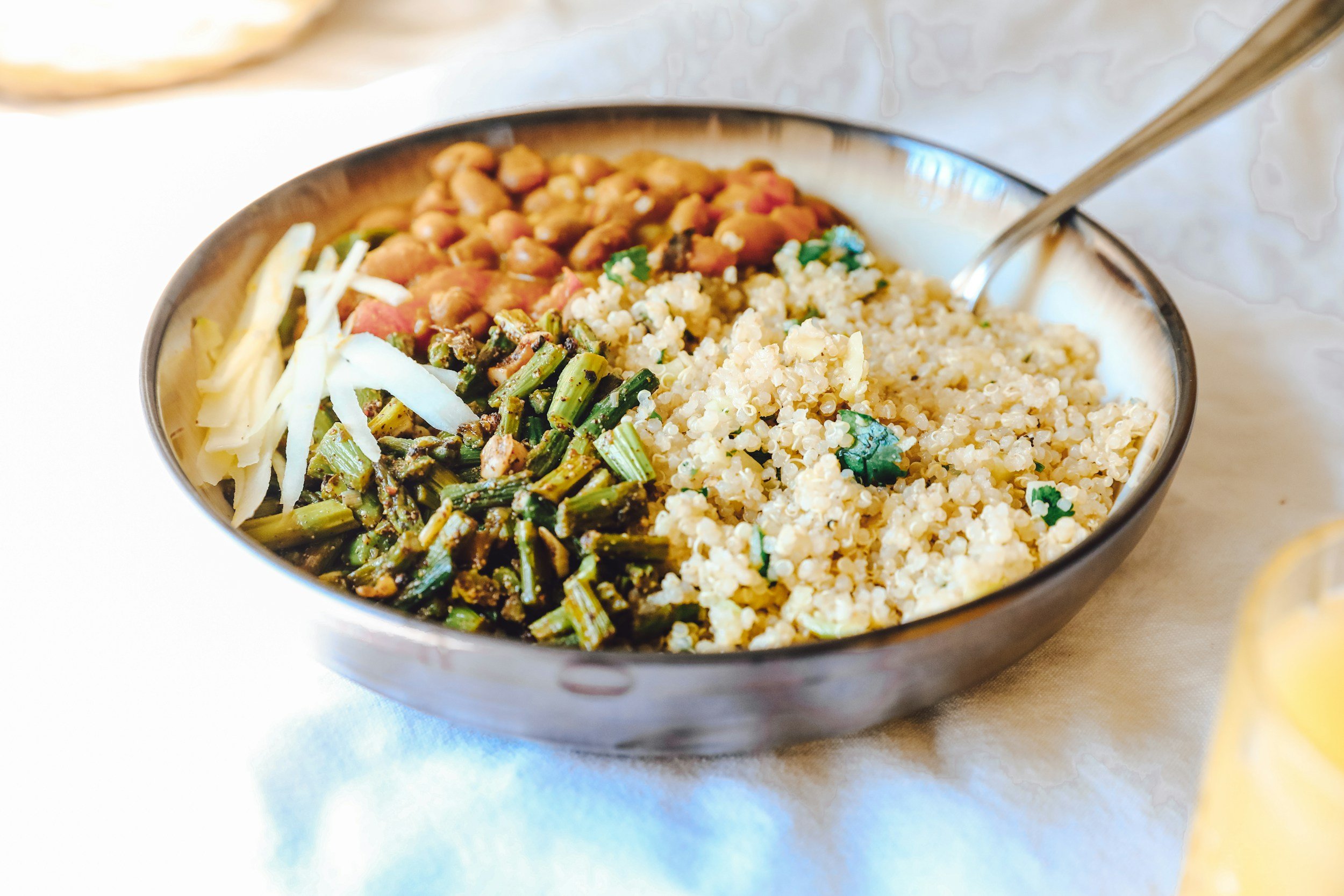 A bowl of assorted vegetables, grains, and beans, including chopped green beans, shredded cabbage, cooked chickpeas, and cooked quinoa, served on a light-colored table.