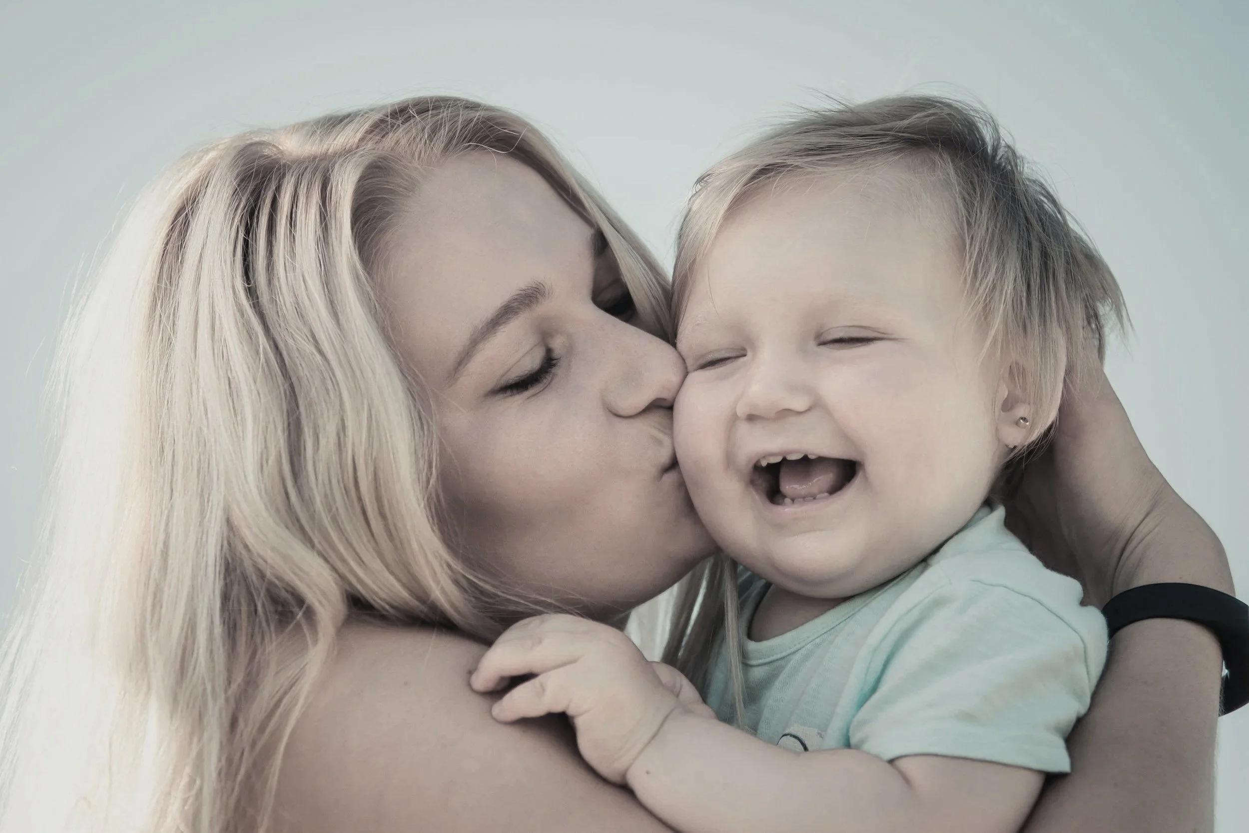 A woman with long blonde hair kissing a smiling young child with short blonde hair on the cheek.