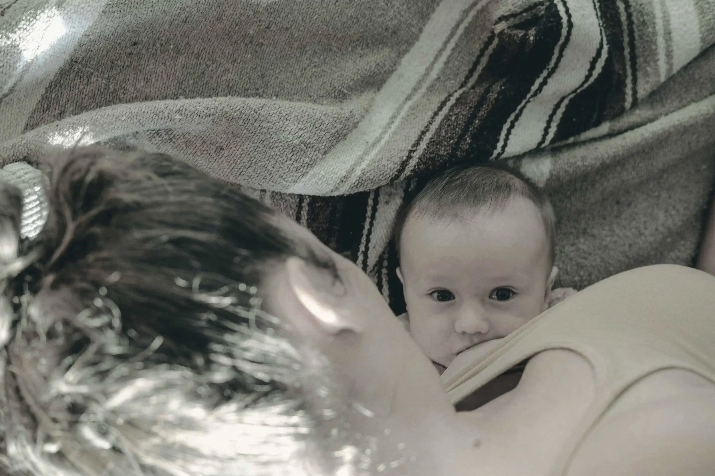 A baby looking at the camera while lying on a person's chest, with an adult person with dark hair and a towel in the background.