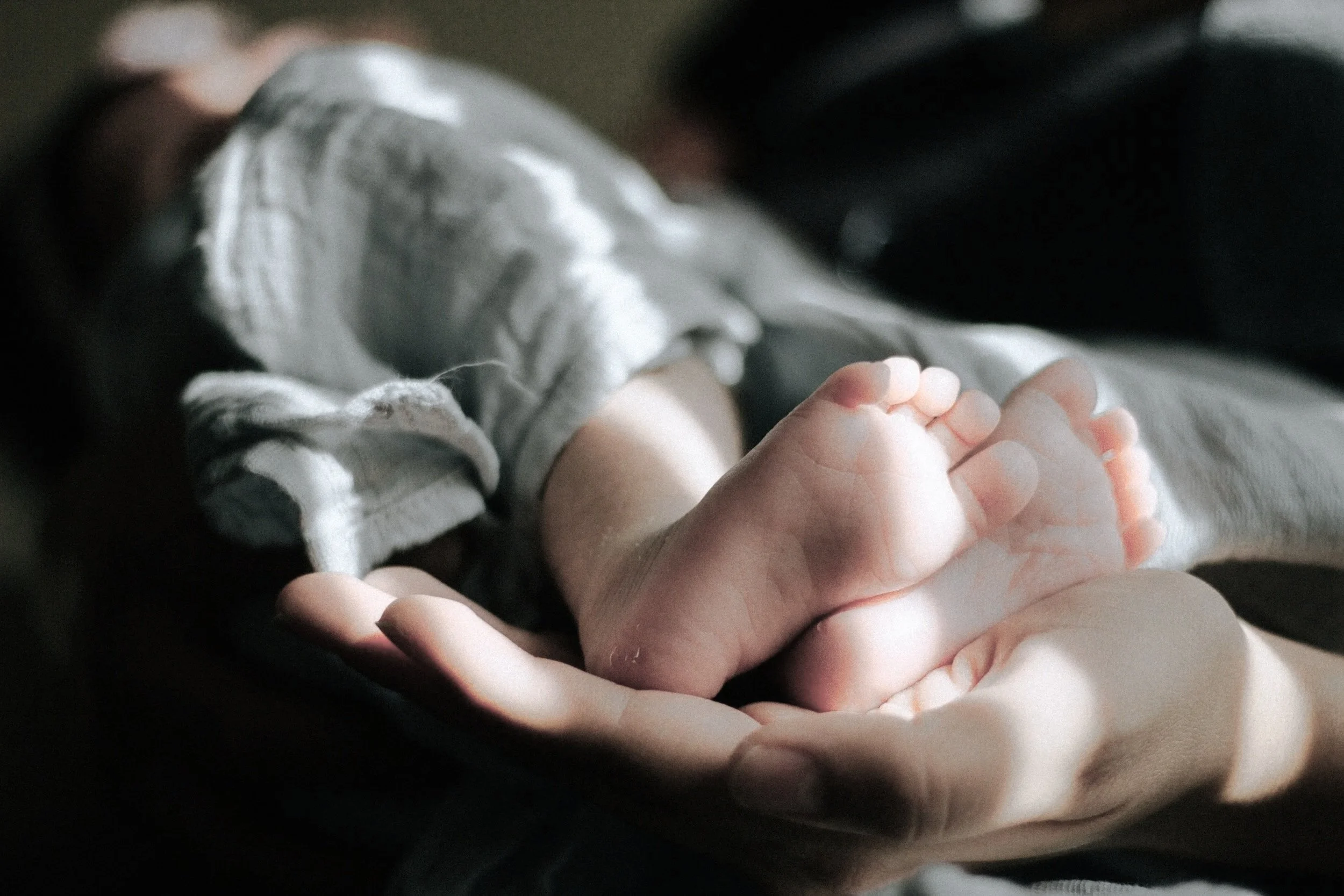 Close-up of a small baby's hand holding an adult's finger, with the baby lying on a person's lap wearing light-colored clothing.