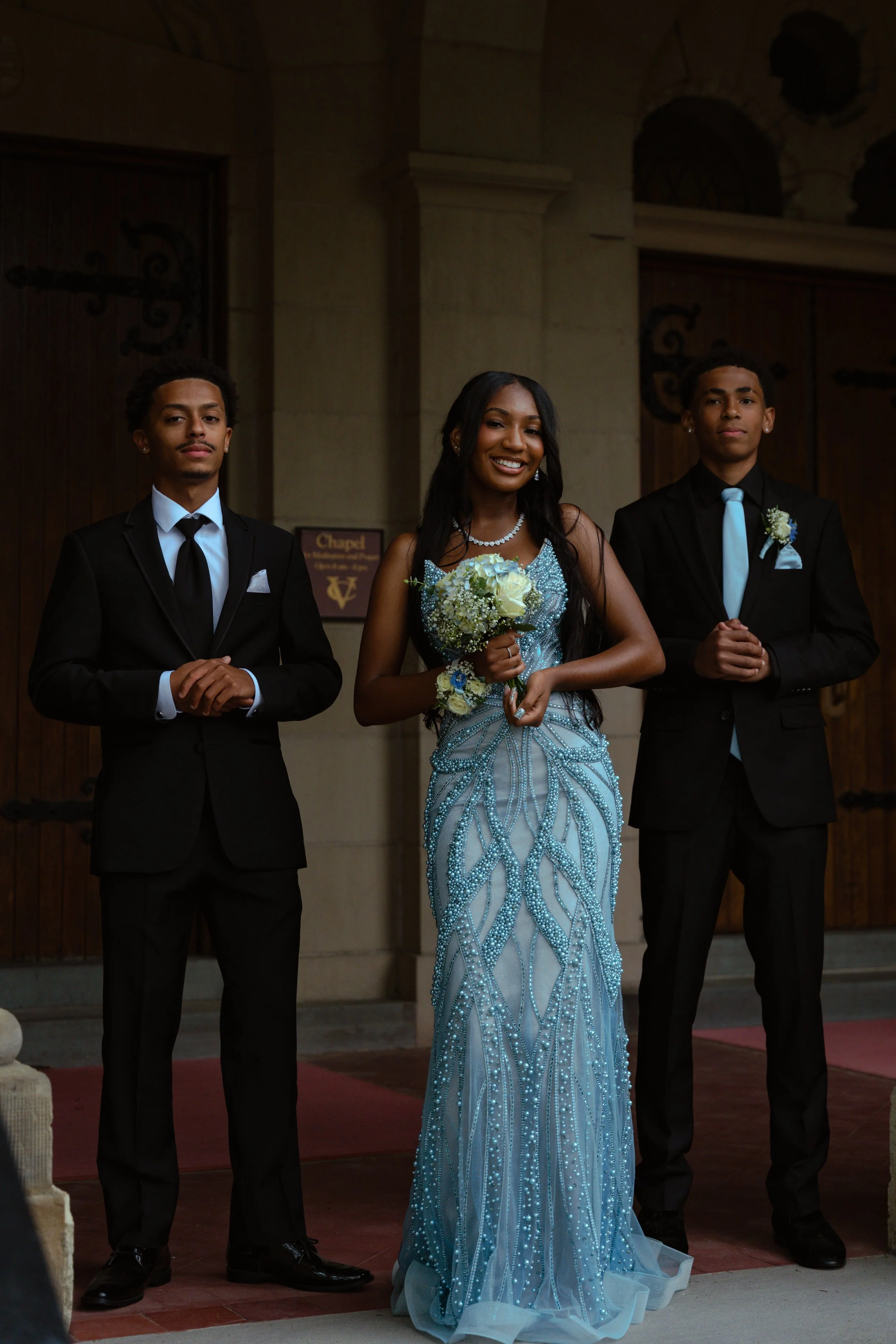 Three young people dressed in formal attire standing in front of a church entrance. The woman in the middle is holding a bouquet and wearing a glittering blue gown, flanked by two young men in black suits with light blue ties and boutonnieres.