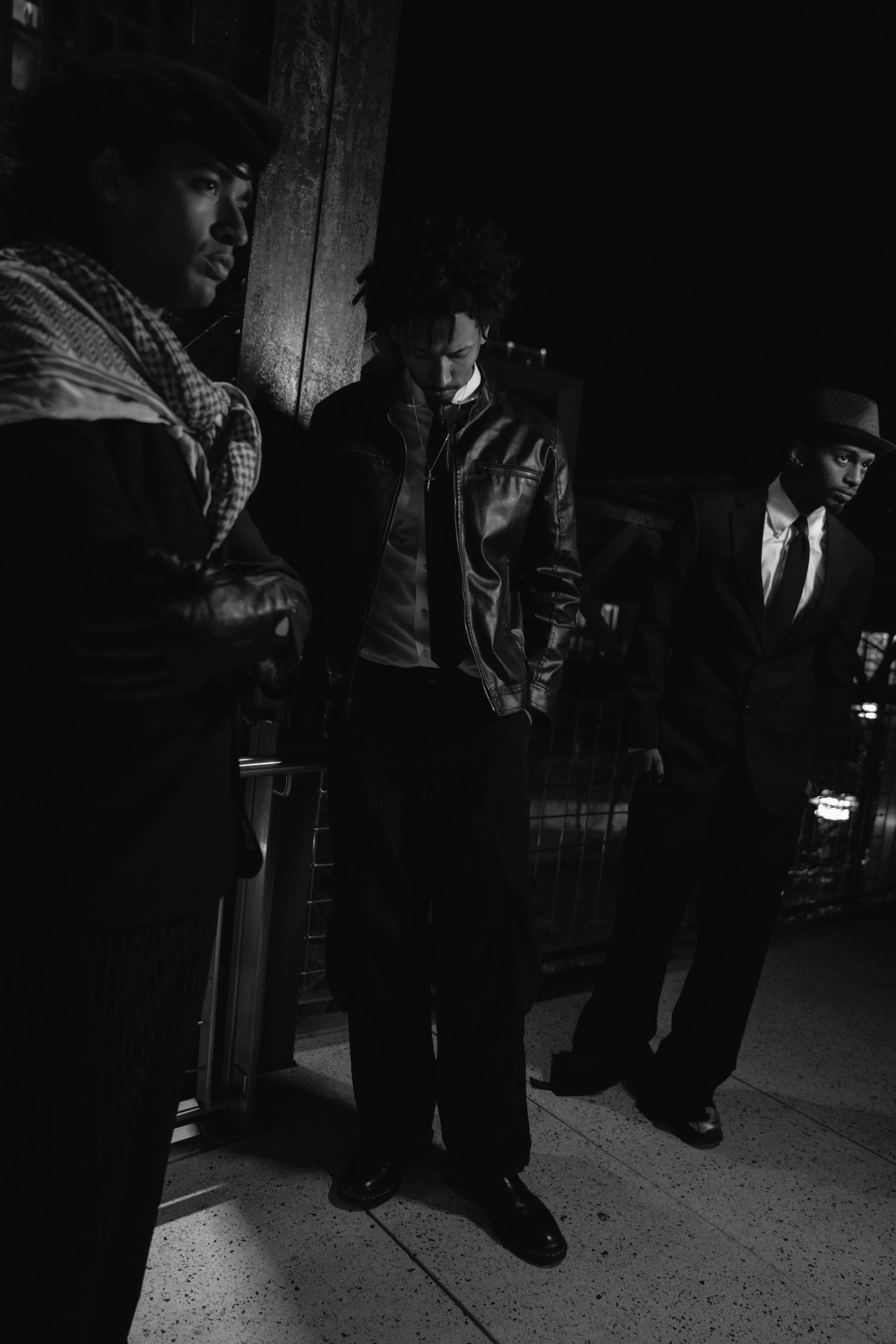 Three men standing on a sidewalk at night, dressed in formal and casual attire, leaning against a wall and looking down or to the side.