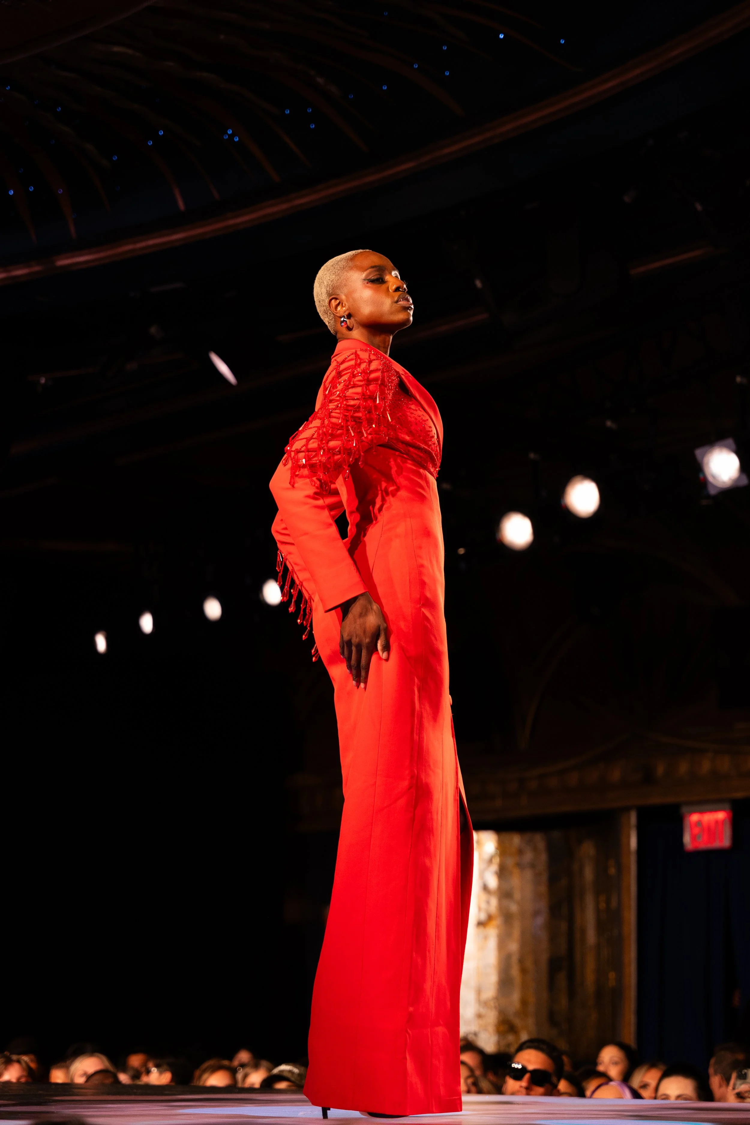 A woman with blonde hair styled in a short buzz cut walking on a runway wearing a red outfit with detailed shoulders, in an indoor venue with an audience and hanging lights.